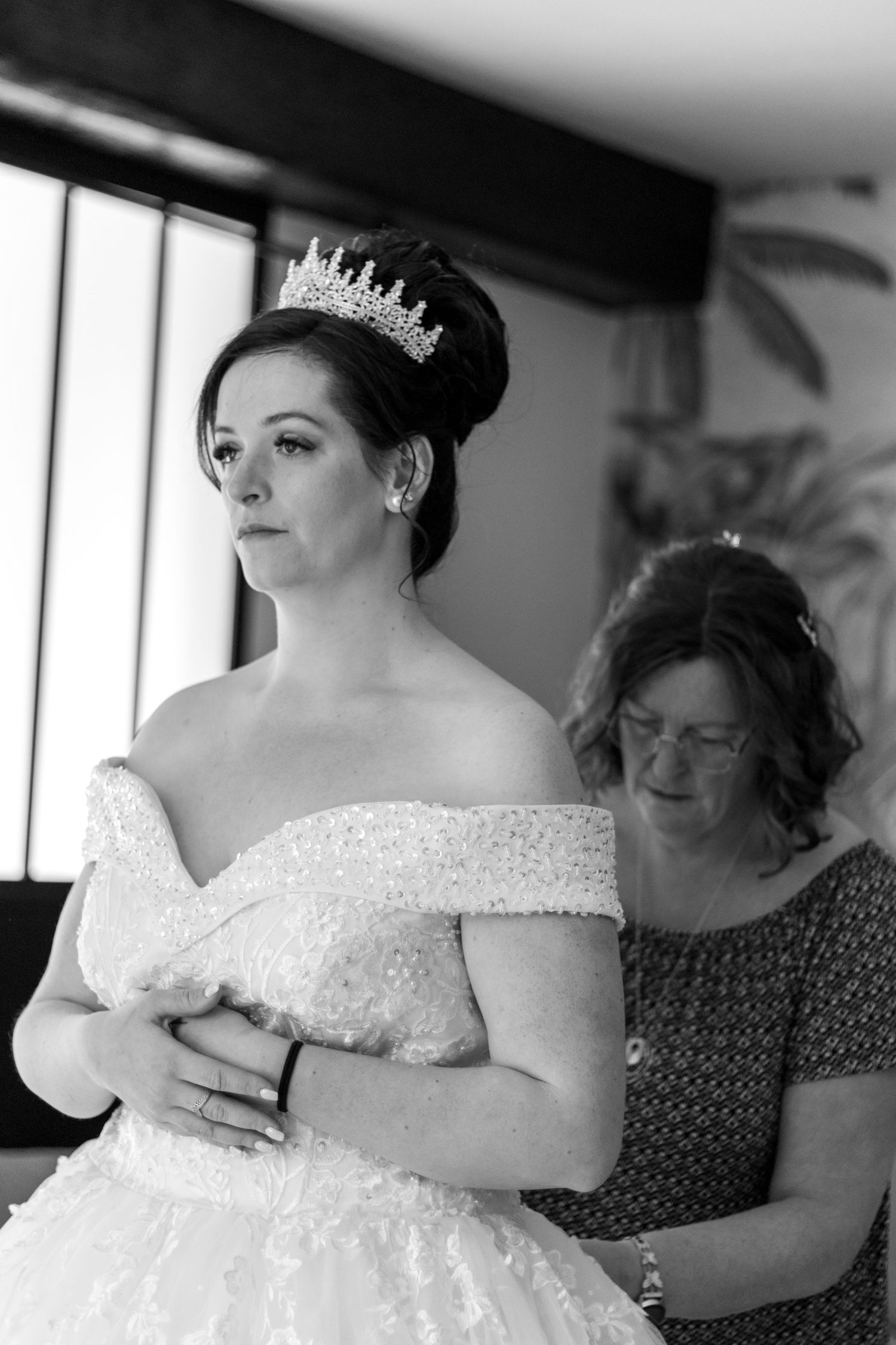 Black and white photo of a bride in an off-shoulder gown and tiara being helped by an older woman.