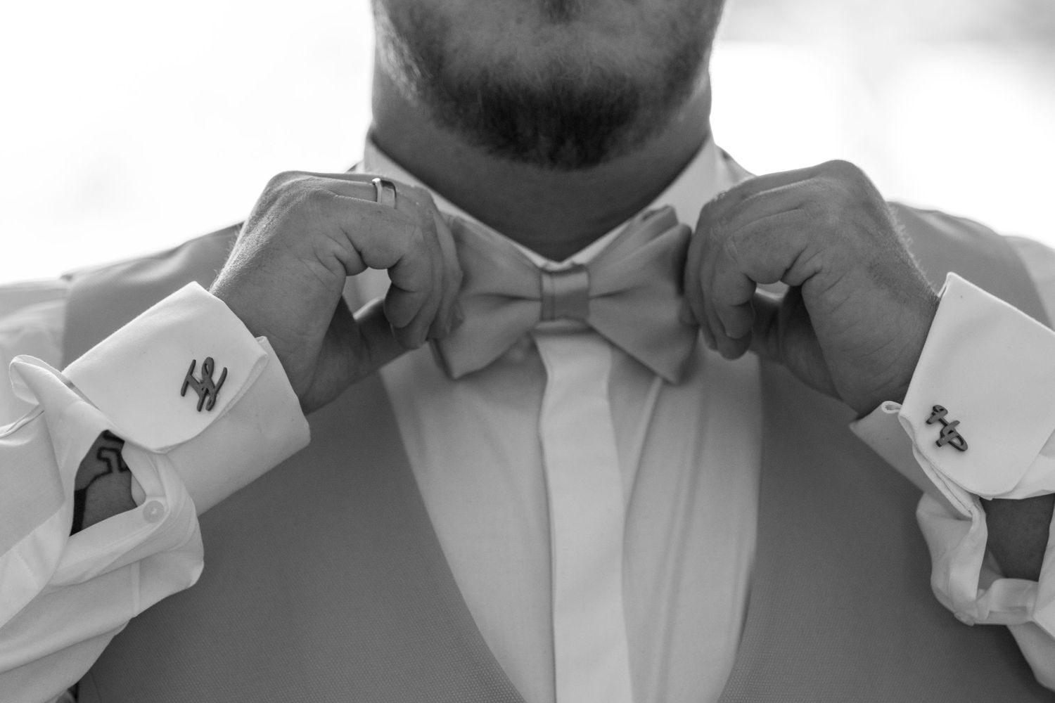 Black and white photo of a groom adjusting his bow tie, wearing a suit with monogrammed cufflinks.