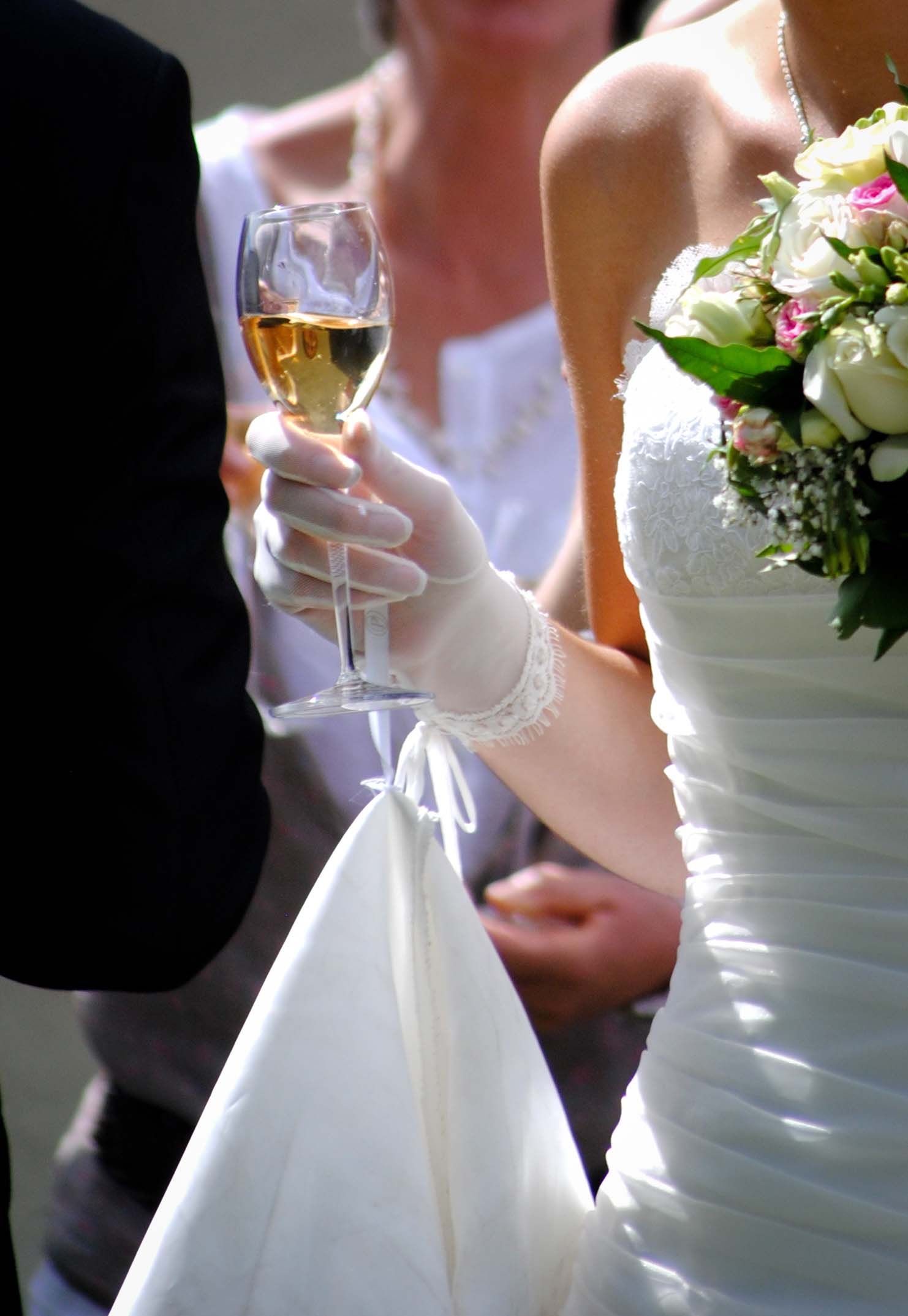 Bride in white gown and gloves holding champagne glass with bouquet at wedding celebration.