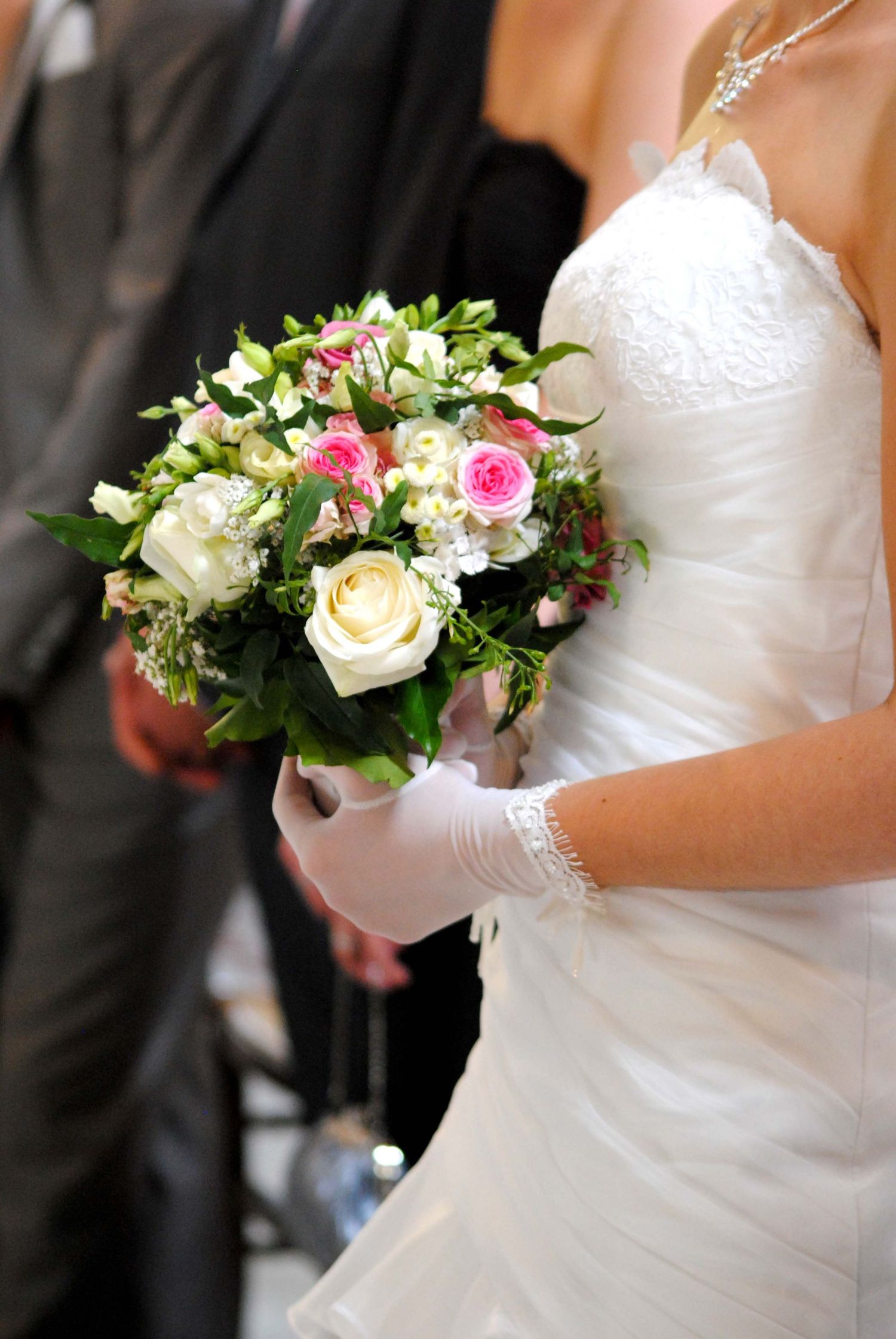 Bride in white strapless gown wearing lace gloves holding a bouquet of pink and white roses at a wedding.