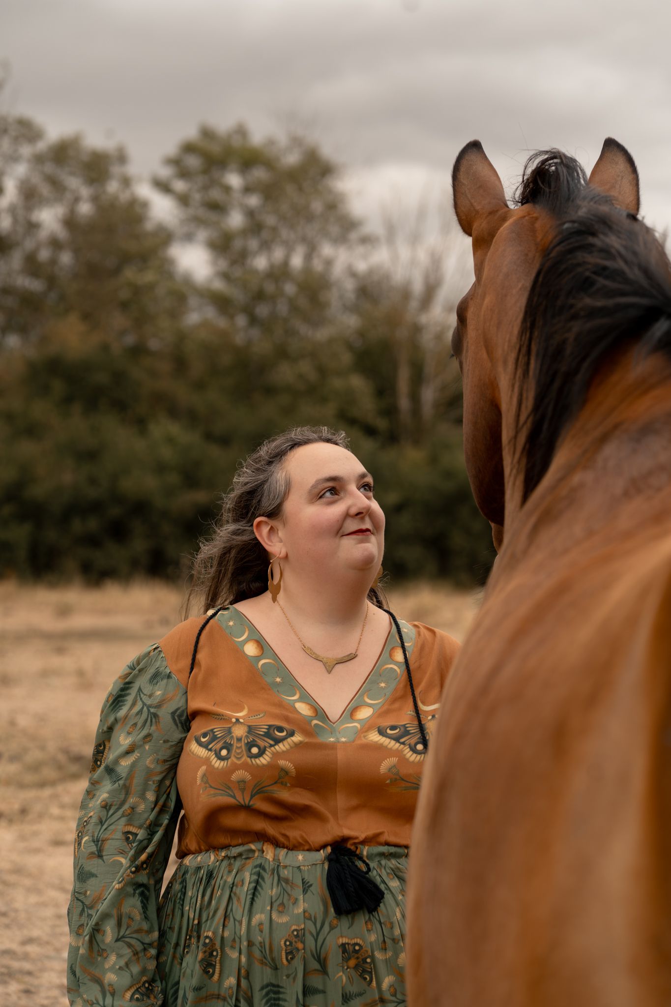Woman in bohemian dress smiling at a brown horse in a rural field under cloudy skies.