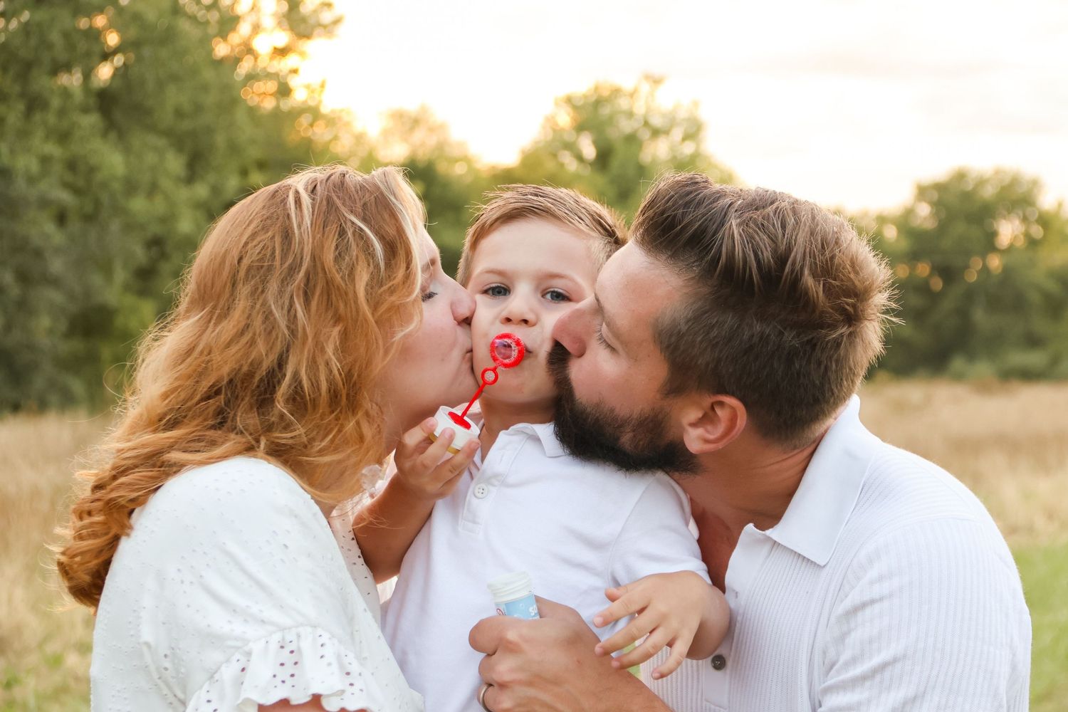 Parents kissing their toddler son holding a lollipop in a sunny outdoor field.