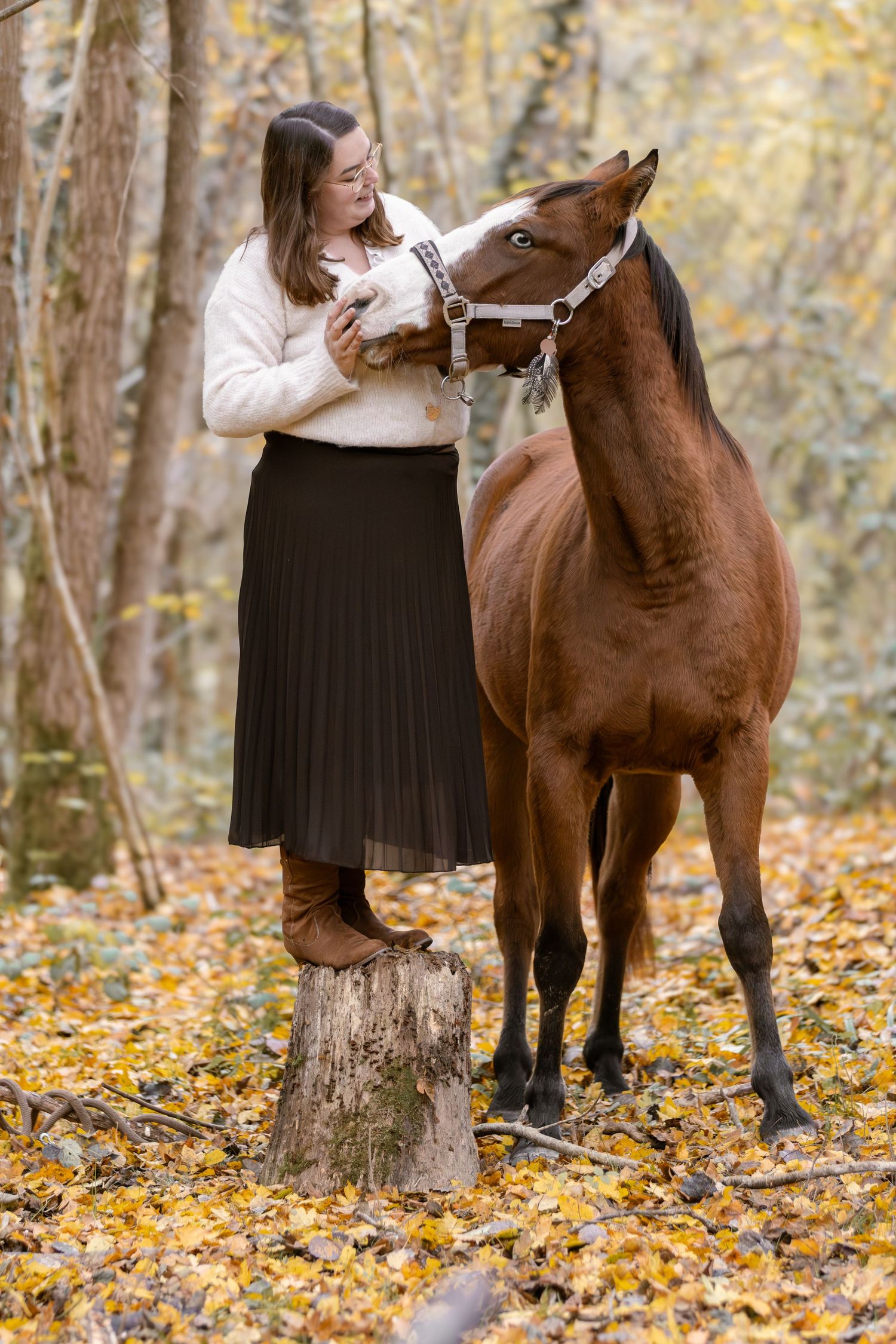 Woman in cream sweater and dark skirt bonding with a brown horse in fall forest.