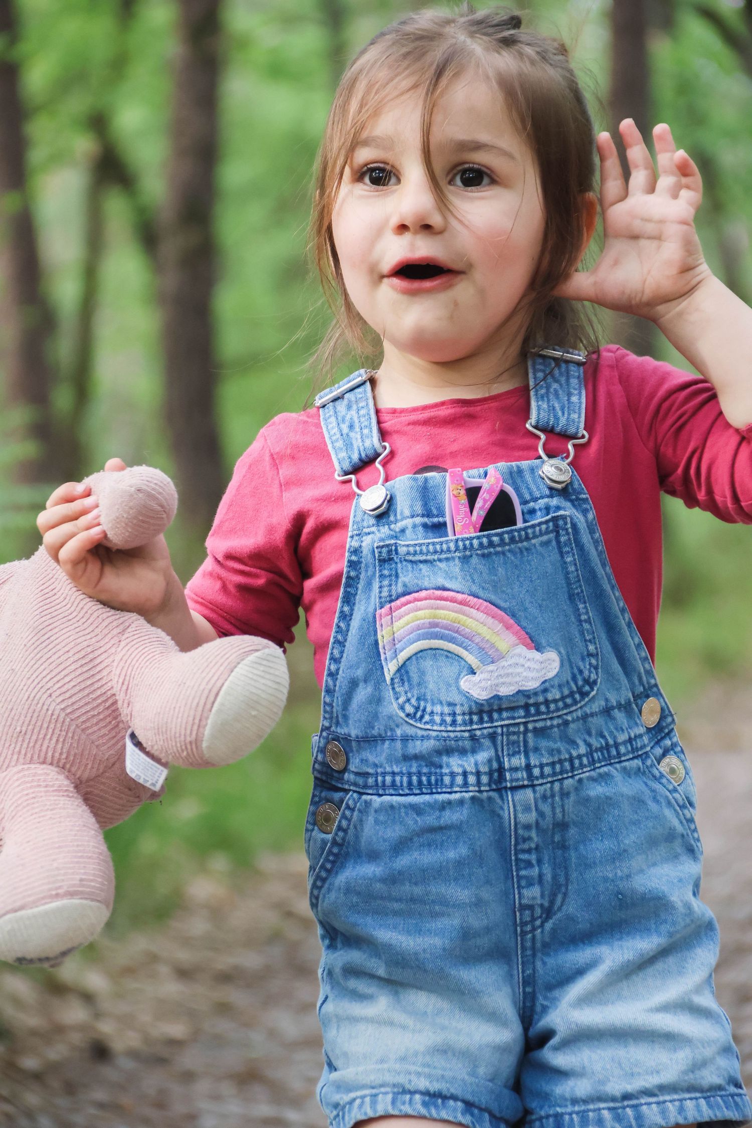 Séance famille lifestyle — petite fille heureuse en salopette jean tenant un doudou rose dans une forêt verdoyante, photographe Soissons