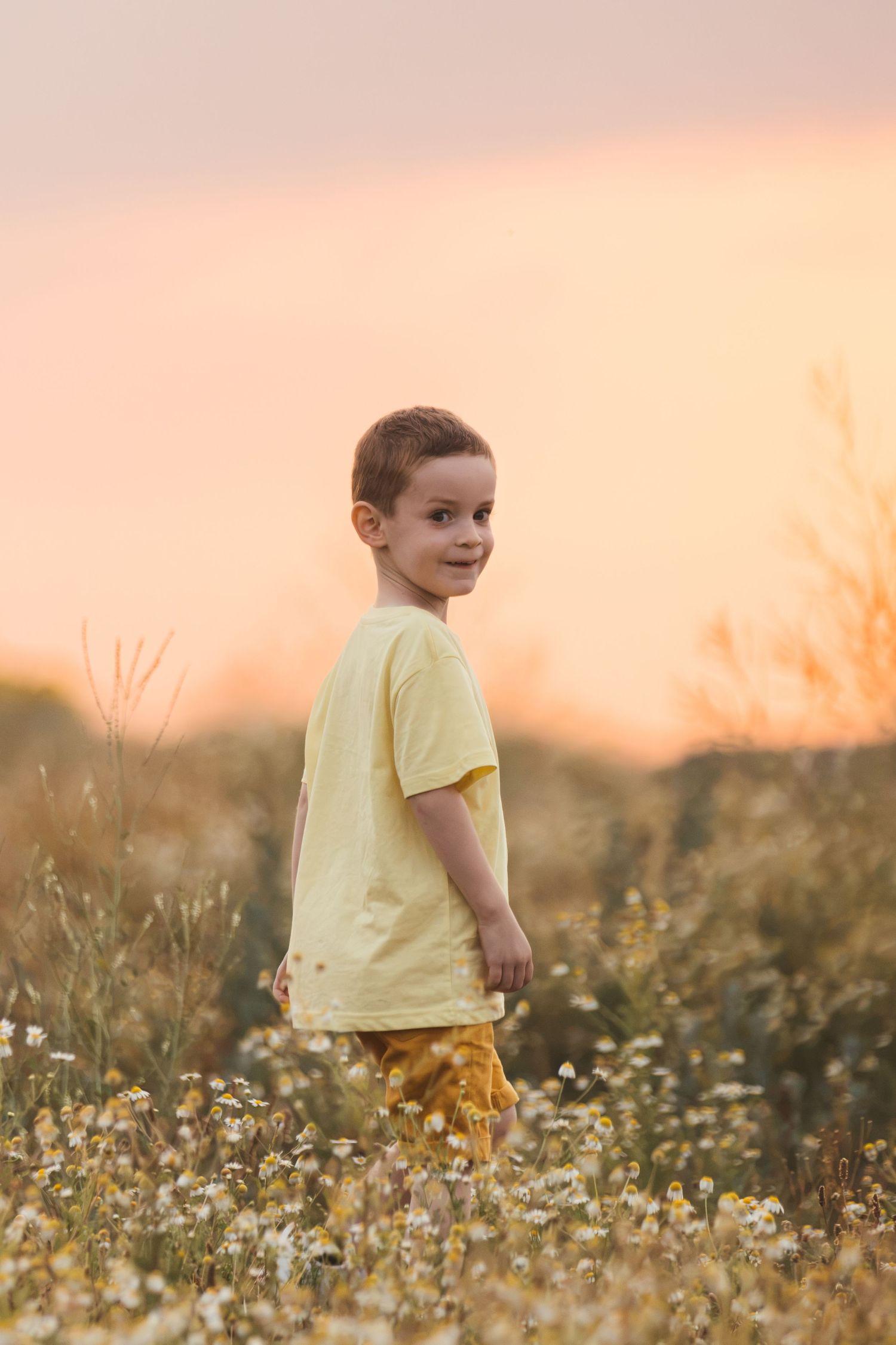 Young boy in yellow shirt standing in wildflower field at golden sunset, smiling over his shoulder.
