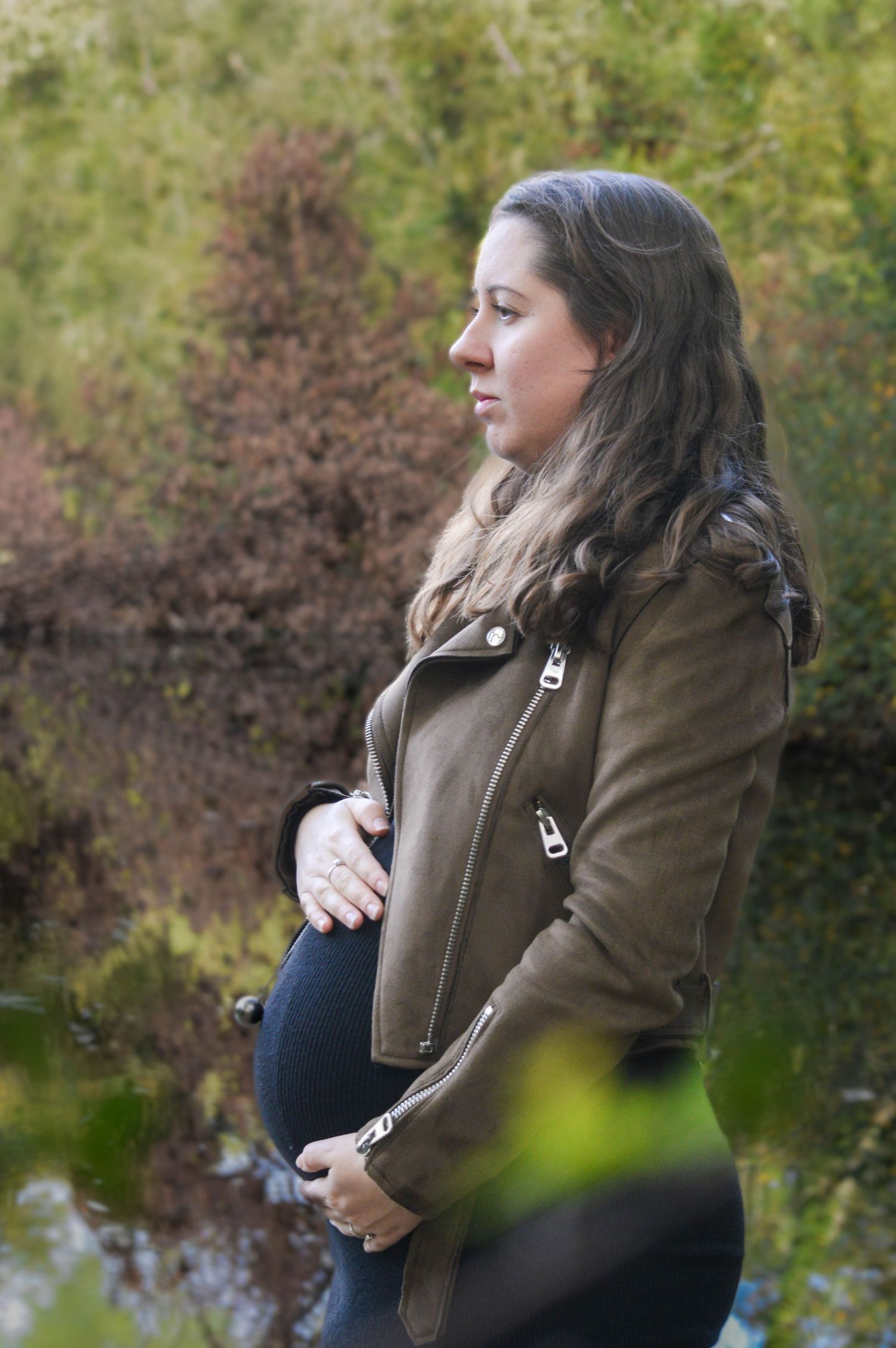 Séance grossesse lifestyle — femme enceinte en veste kaki tenant son ventre au bord d'un étang en forêt, photographe Charleville-Mézières