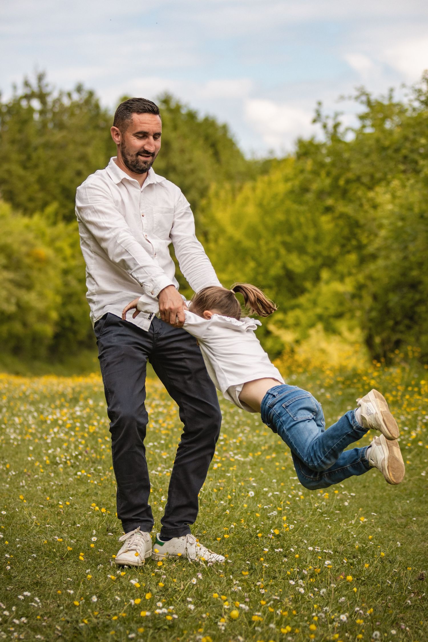 Father swinging his child in a sunny meadow with wildflowers, both wearing white shirts, enjoying outdoor family fun.