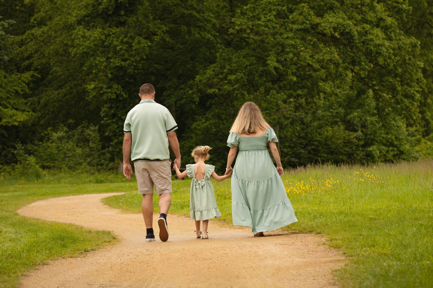 Family of three holding hands walking down a winding path surrounded by lush green trees.