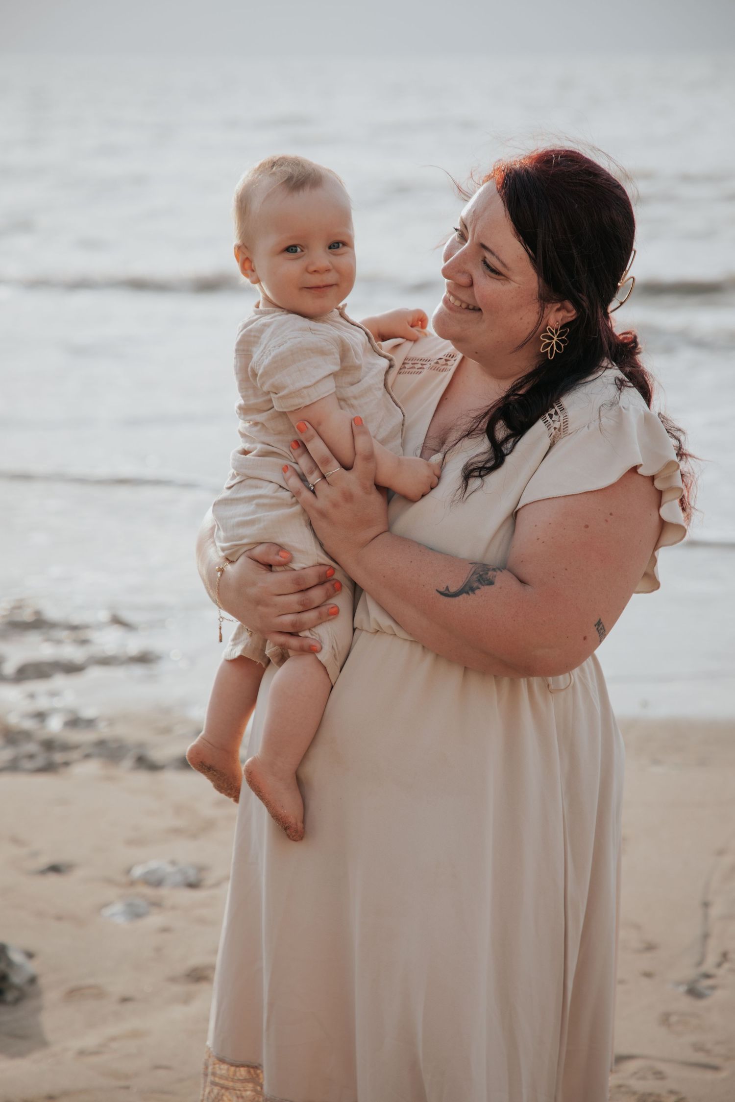 A smiling woman in a white dress holds a happy baby on a sandy beach with ocean waves in the background.