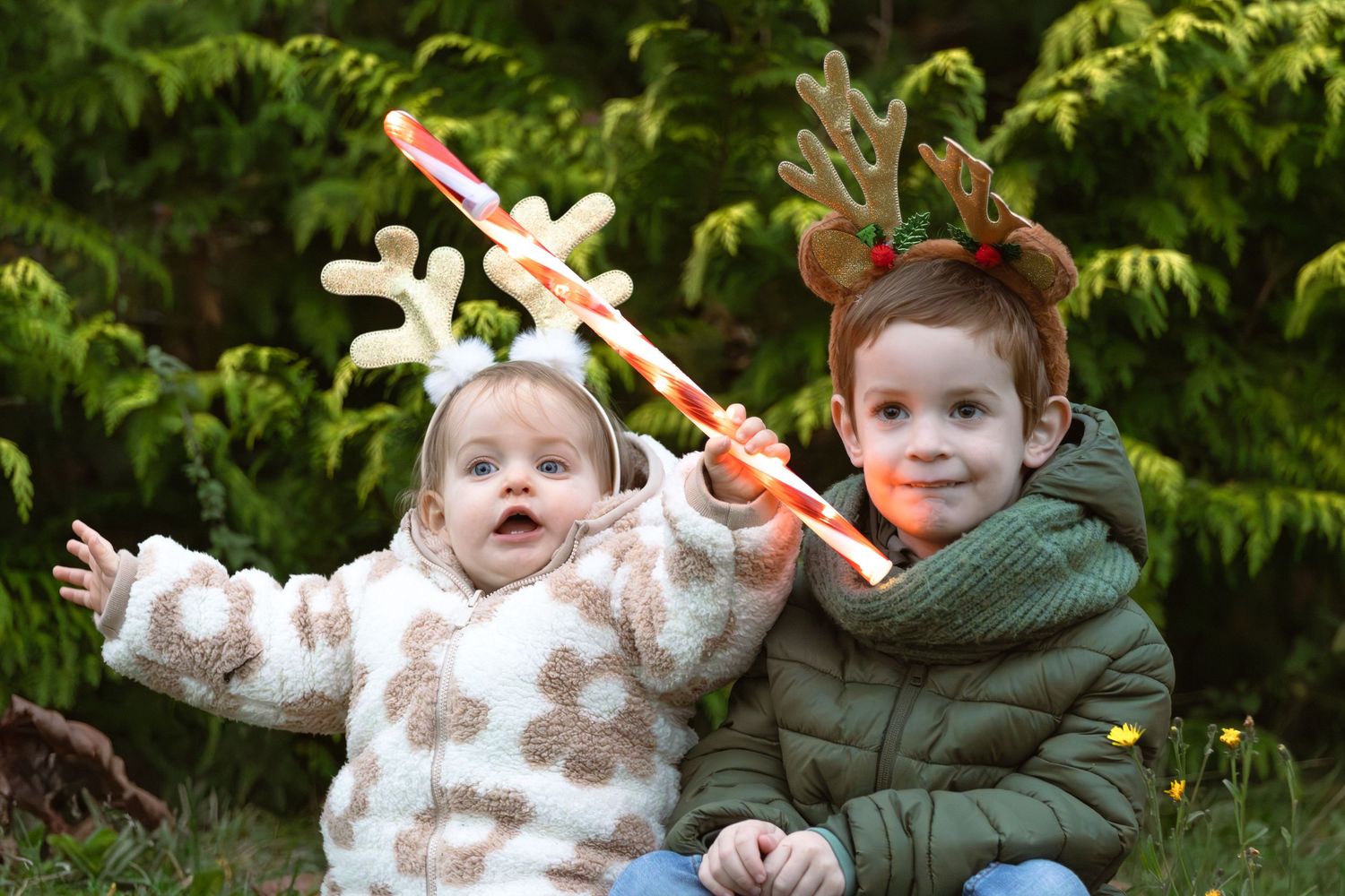 Two young siblings wear reindeer antler headbands outdoors near evergreen trees at Christmas.