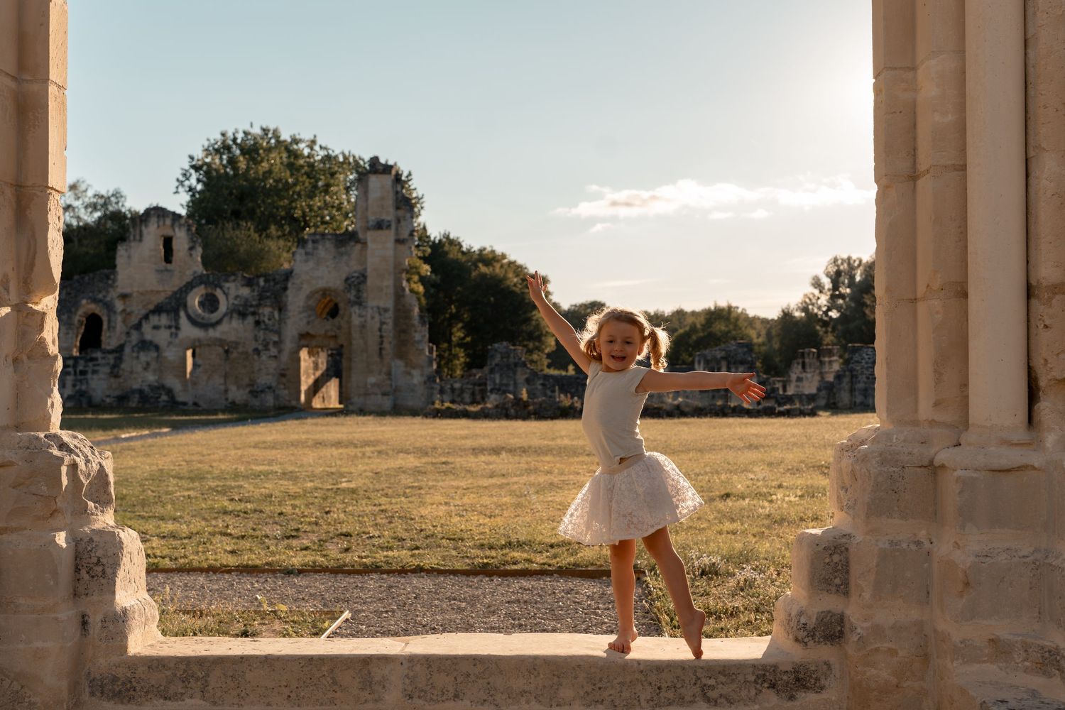 A little girl in a white tutu dress dances gracefully among ancient stone ruins at golden hour.