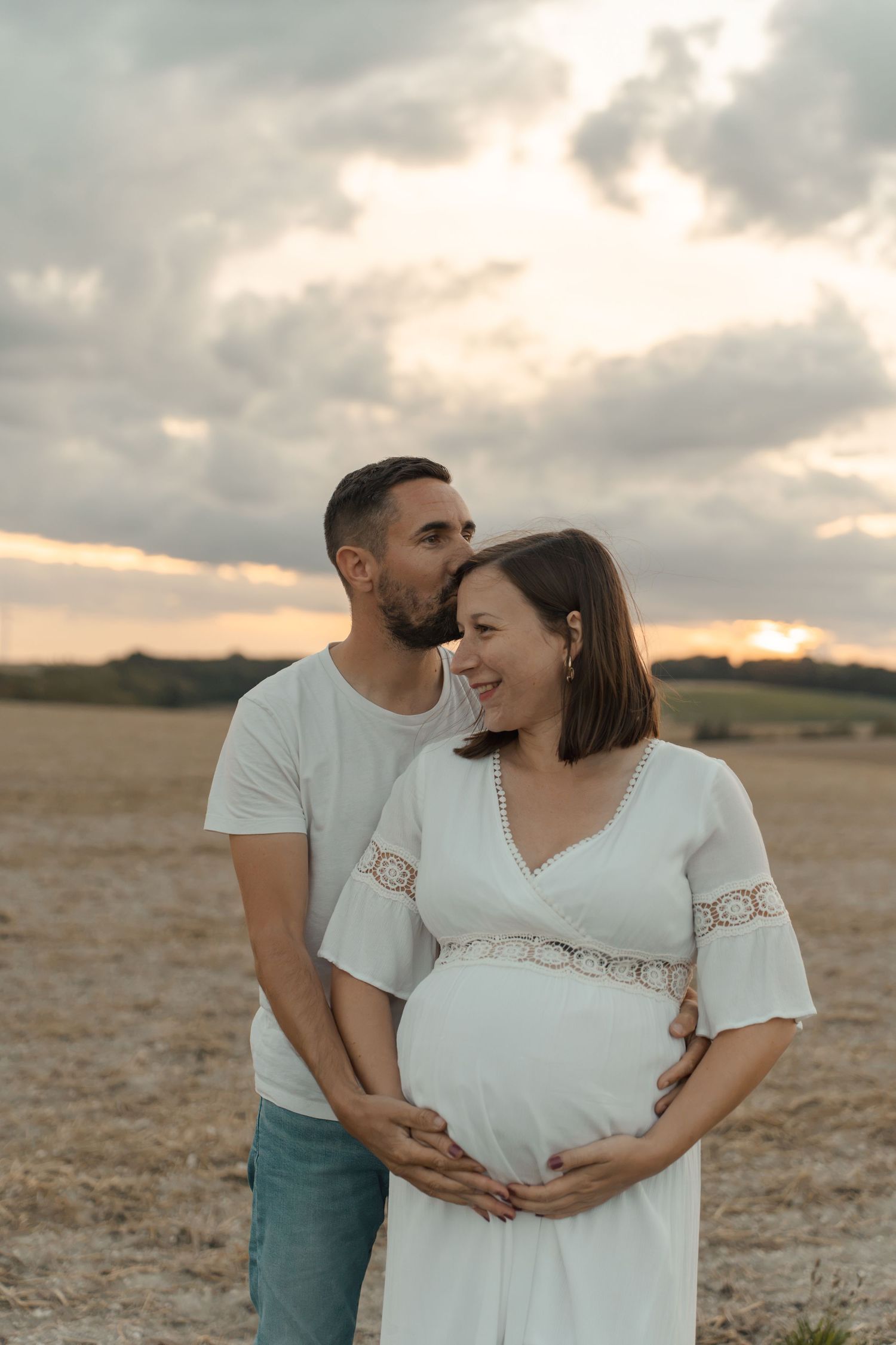 Happy couple in field at sunset, man kissing pregnant womans head while holding her baby bump.