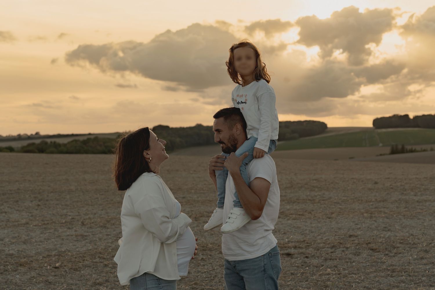 A family of three poses at sunset in a field, with a man holding a child on his shoulders beside a pregnant woman.