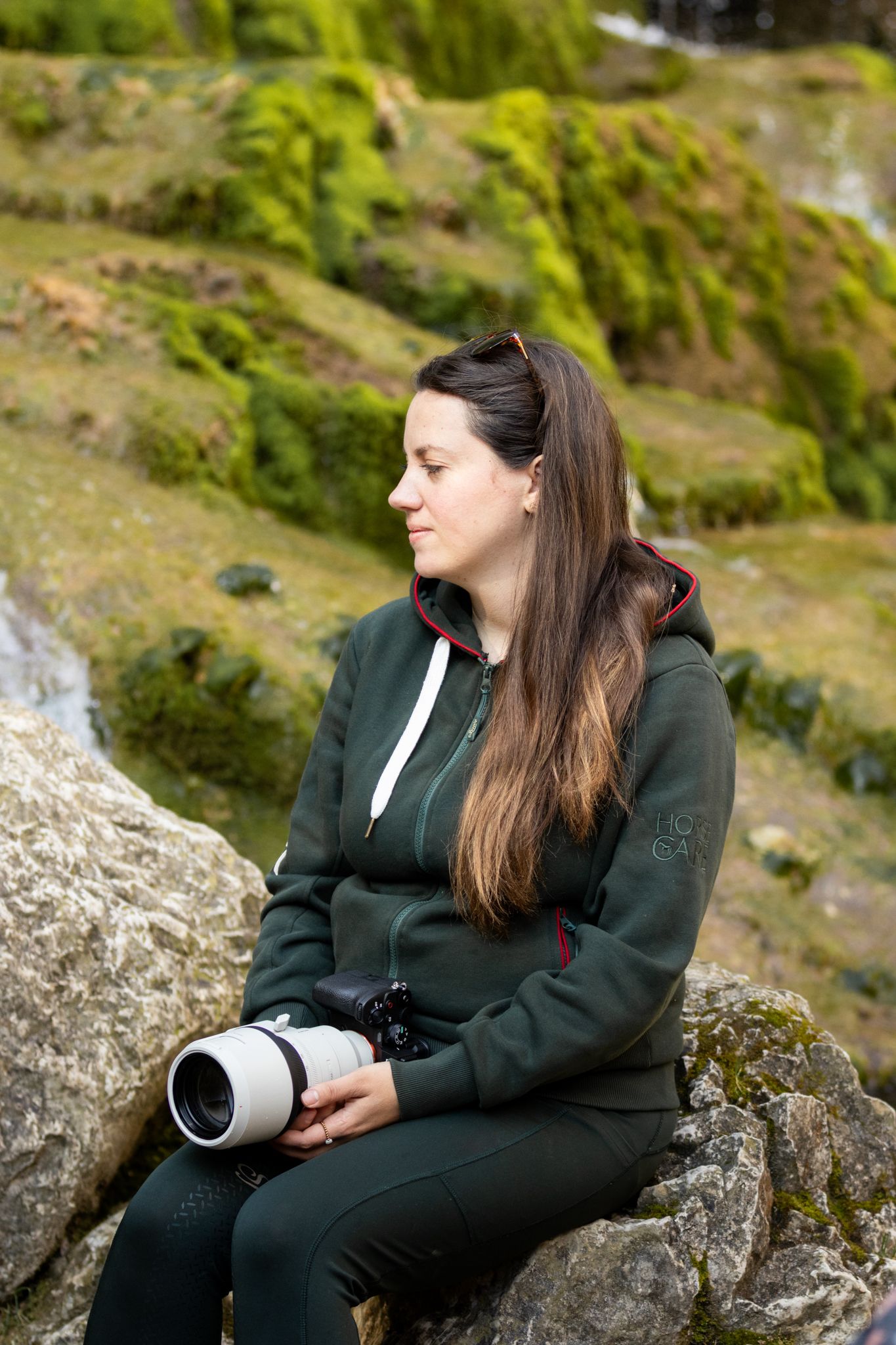 Woman photographer sitting on mossy rocks outdoors, holding a camera with telephoto lens, wearing a green hoodie.