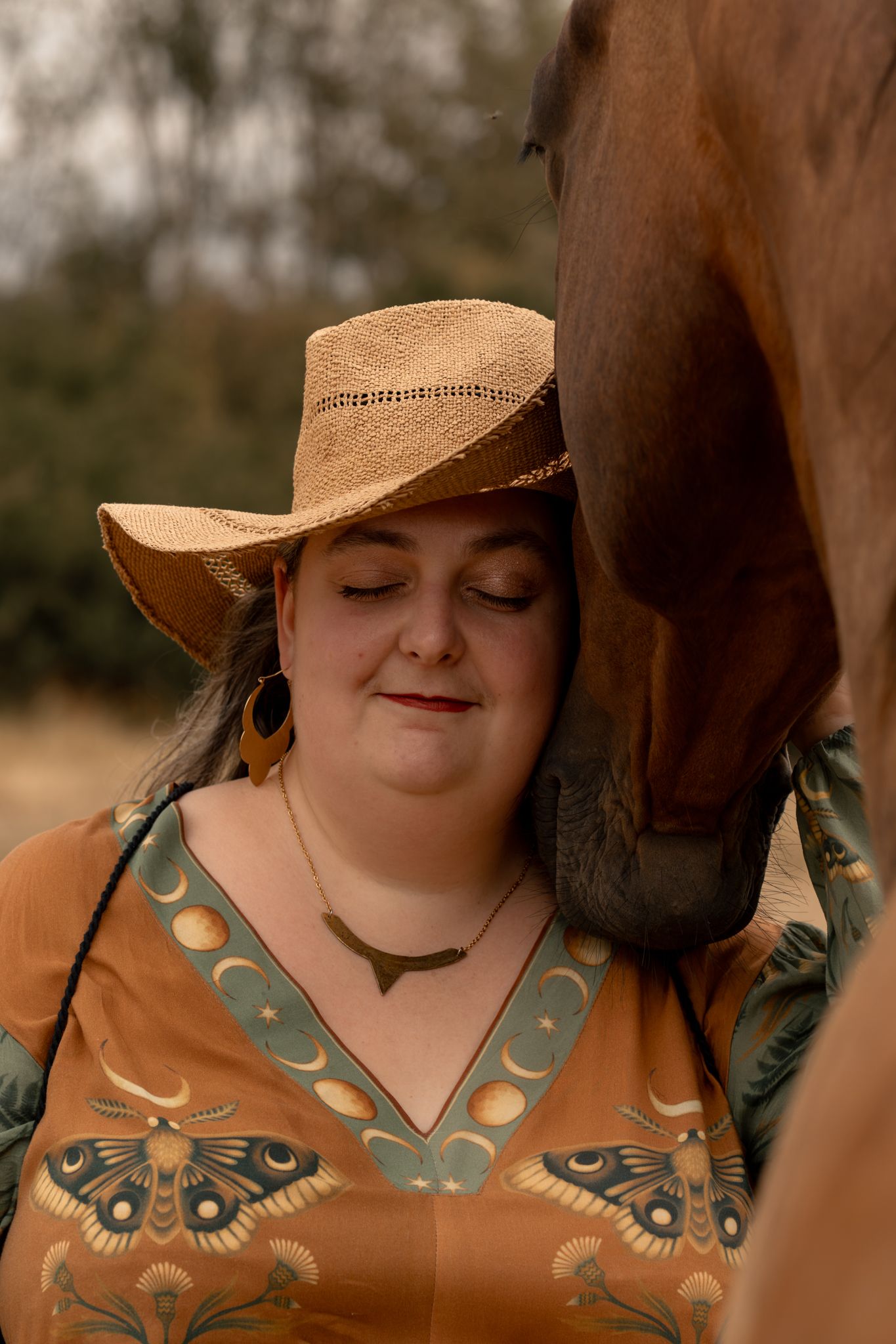 Woman in straw hat with eyes closed enjoying a tender moment with a chestnut horse nuzzling her.