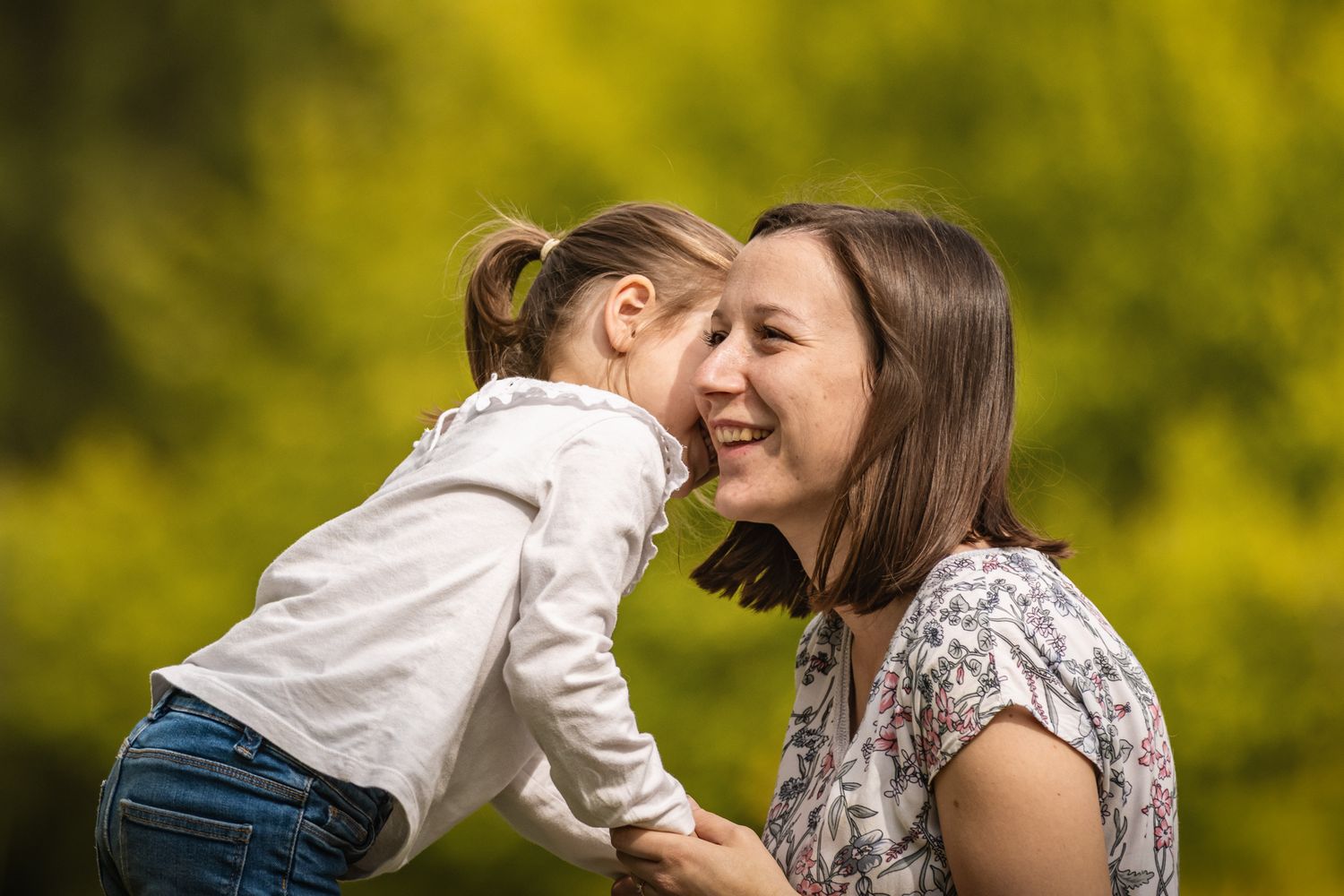 A mother and young daughter share a joyful moment nose-to-nose outdoors with green foliage background.