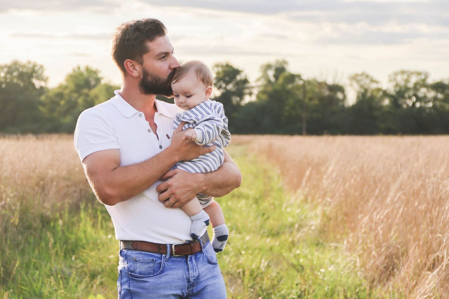 Séance famille — papa barbu en polo blanc berçant son nourrisson en body rayé dans un champ de blé ensoleillé, photographe Aisne Guignicourt