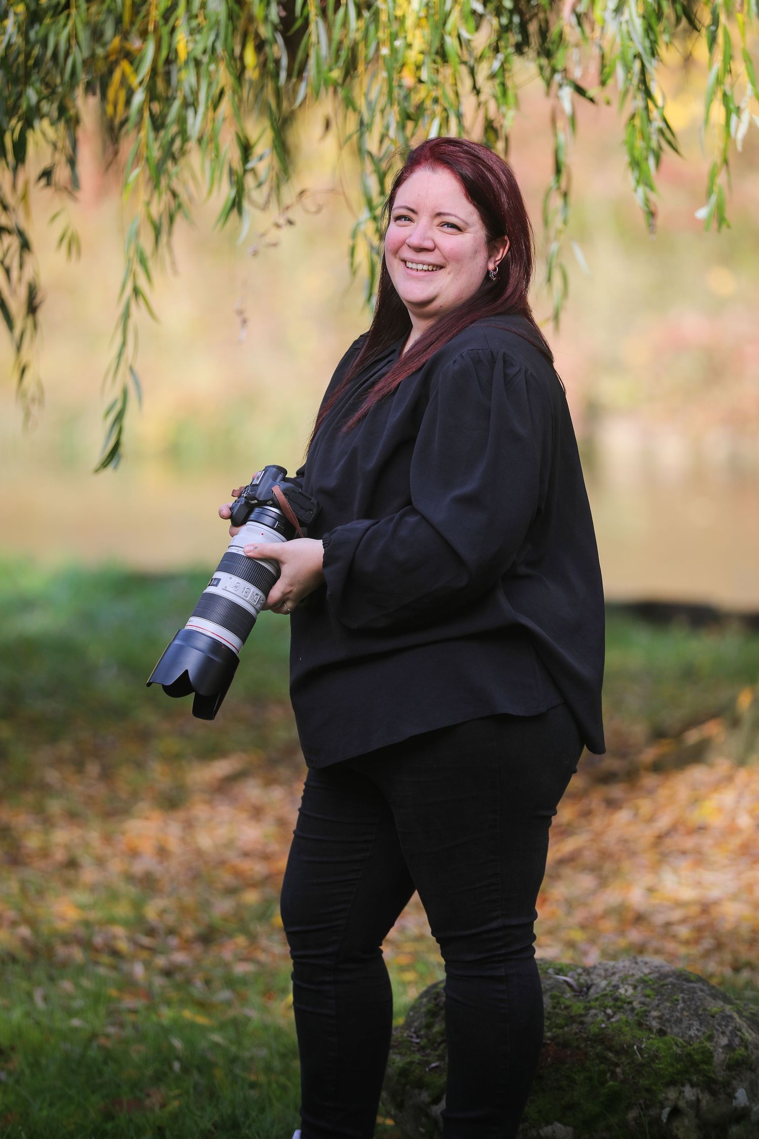 Smiling female photographer holding professional camera with telephoto lens standing under willow tree in fall setting.