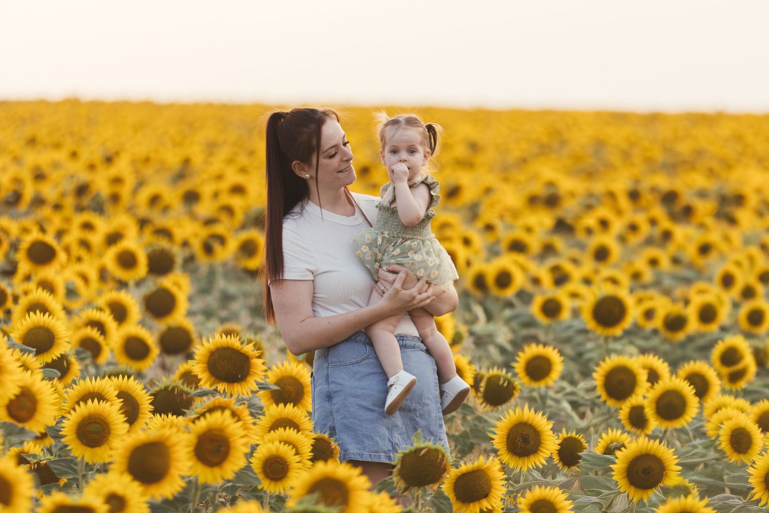 A mother holds her toddler daughter in a vast sunflower field at sunset, both smiling joyfully.