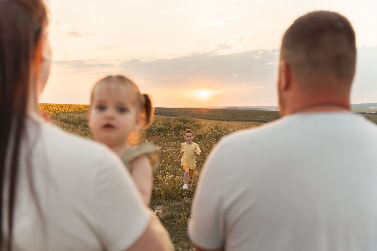 Family at sunset in a field, parents holding baby girl while young child runs toward them.