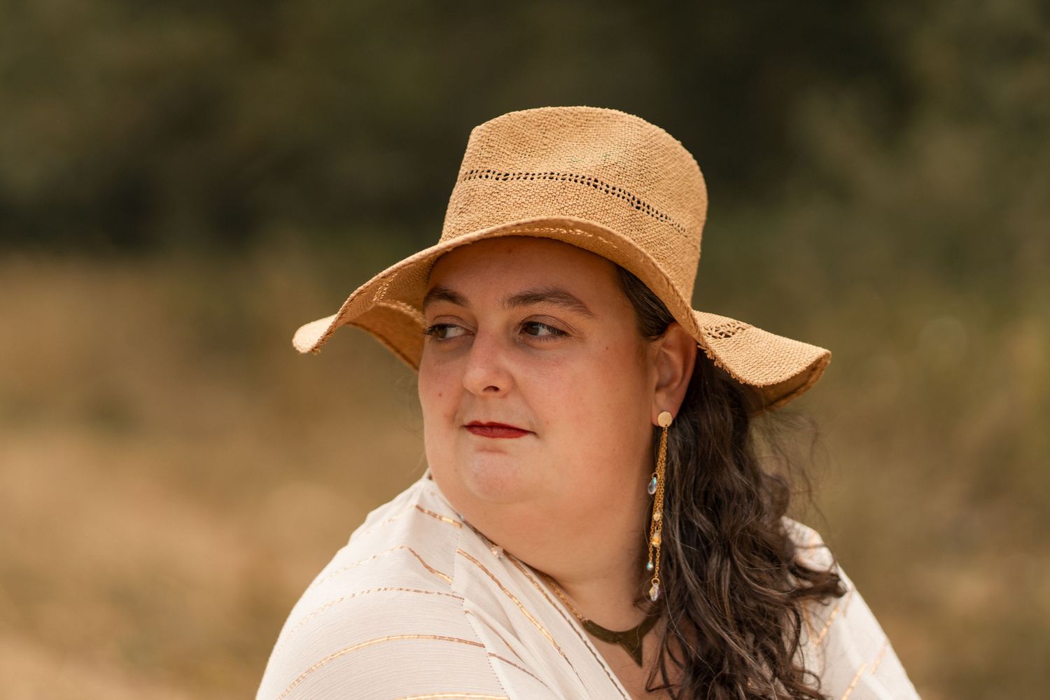 Woman wearing a tan wide-brim hat and white top with dangling earrings, posing outdoors in a natural setting.