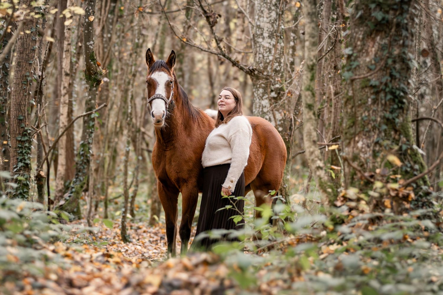 A woman in a cream sweater stands beside a chestnut horse among trees with fallen autumn leaves.