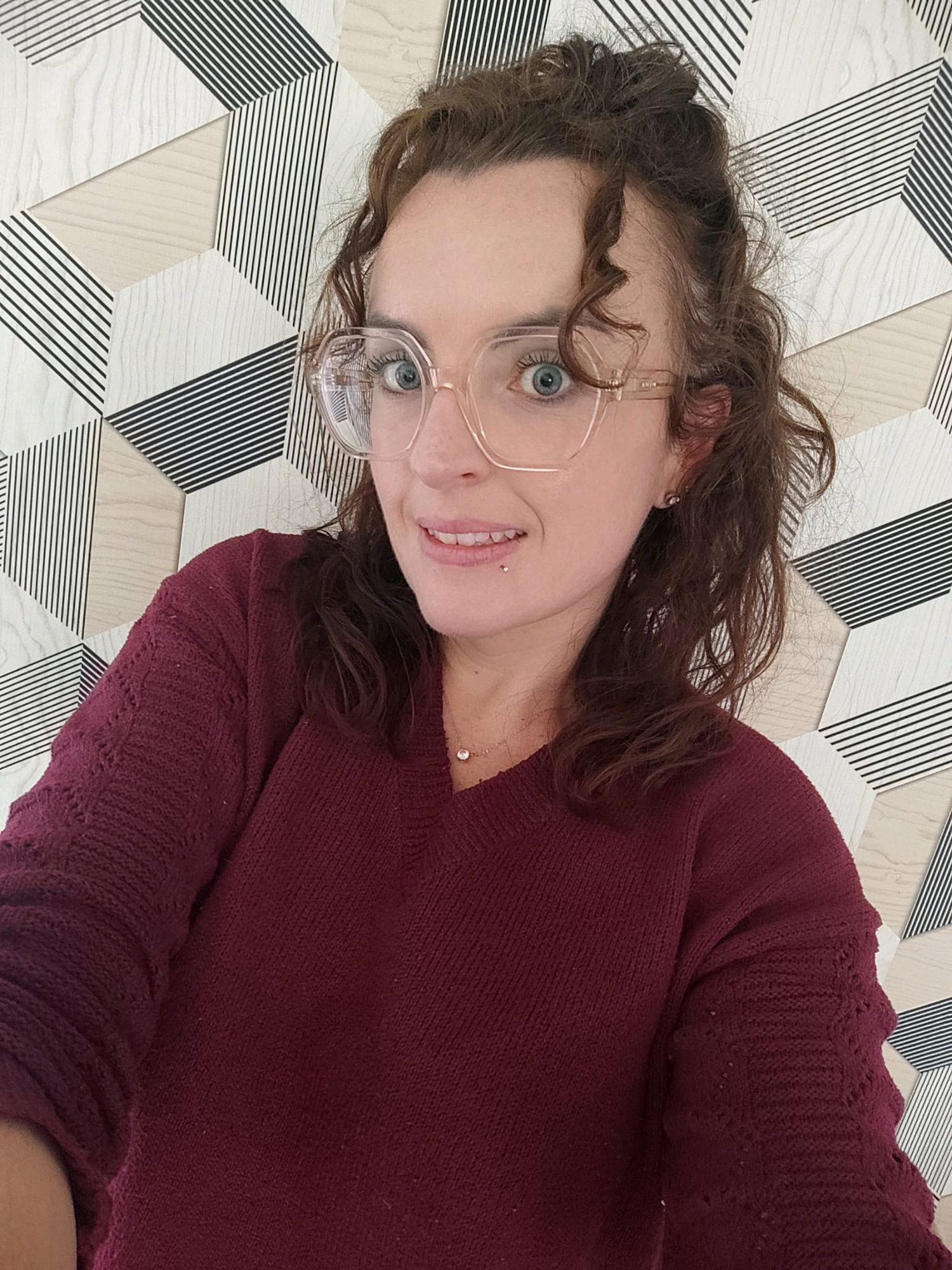 Woman with curly brown hair and clear-frame glasses wearing a burgundy sweater, taking a selfie against a geometric patterned wall.