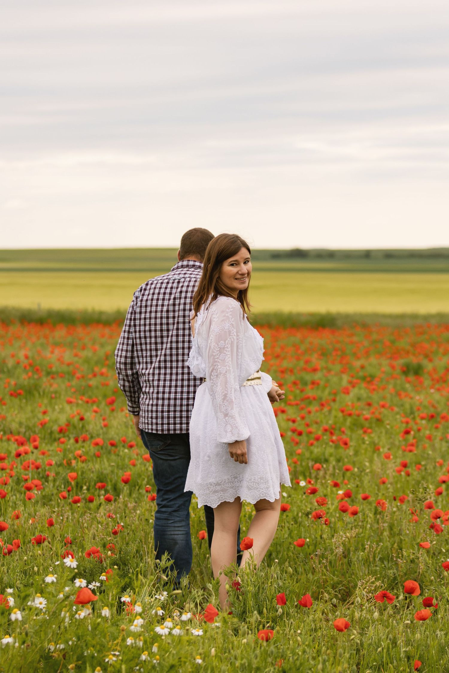 Couple embracing in a vibrant red poppy field, woman in white dress smiling, man in plaid shirt, green meadow background.