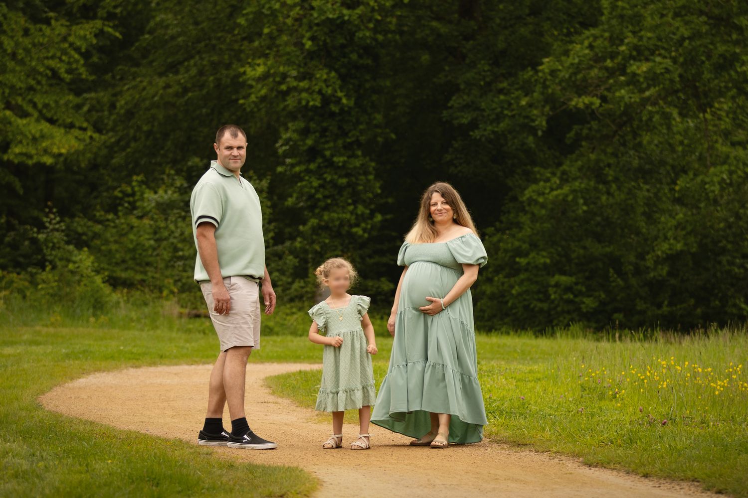 Pregnant woman in green dress with family on park path, husband and toddler daughter, outdoor maternity photo session.