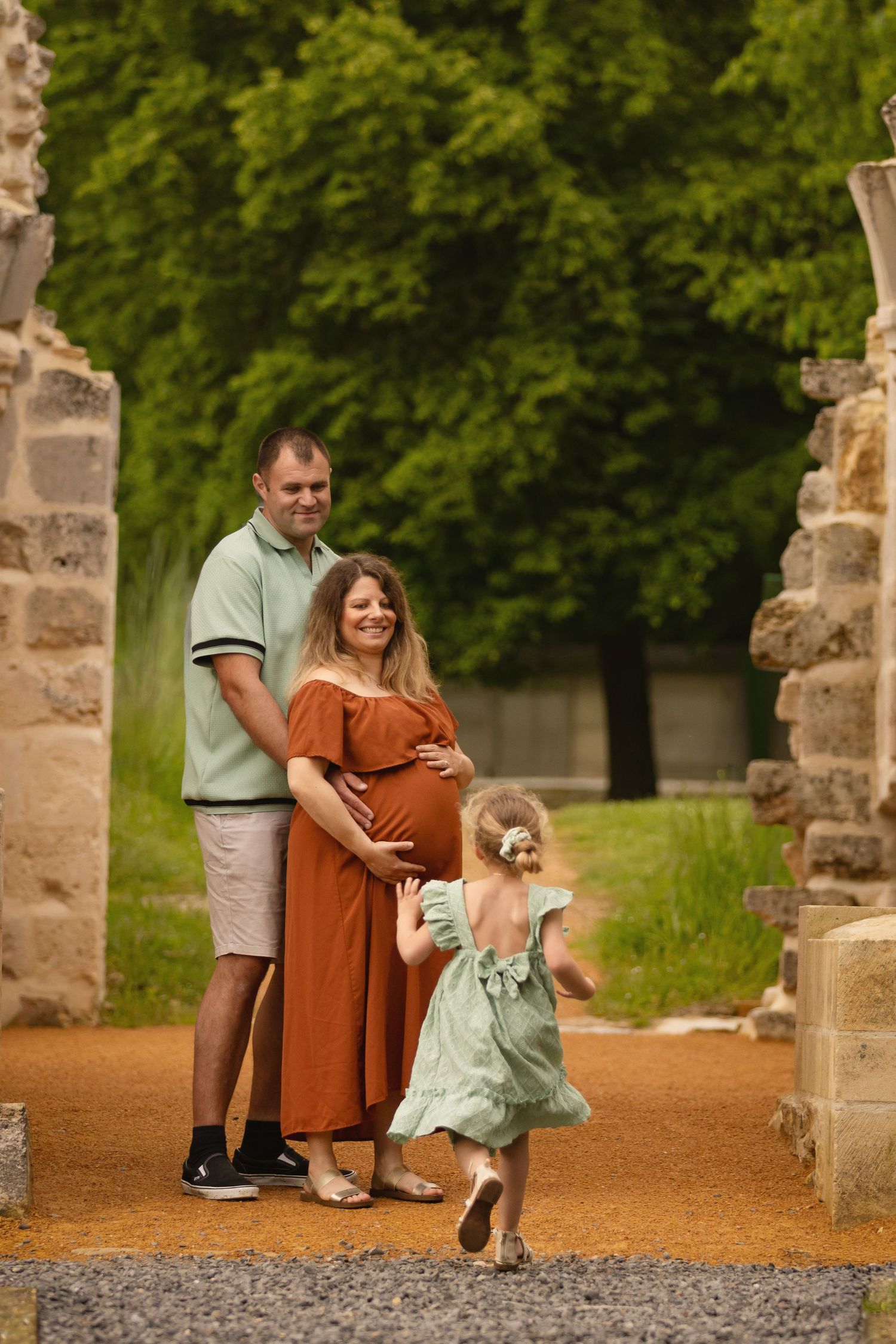 Pregnant woman in rust dress with husband and young daughter in green dress enjoying a family photo outdoors.