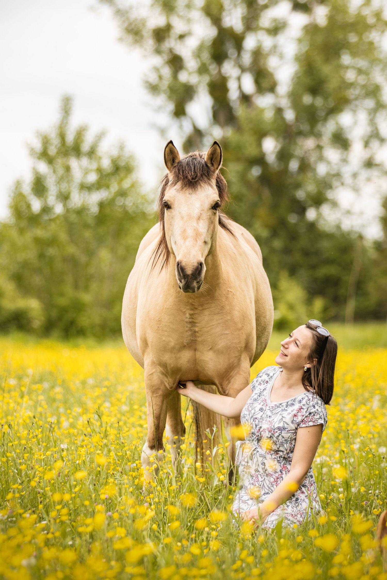 Woman in floral dress kneeling beside a buckskin horse in a vibrant yellow wildflower meadow with green trees behind.