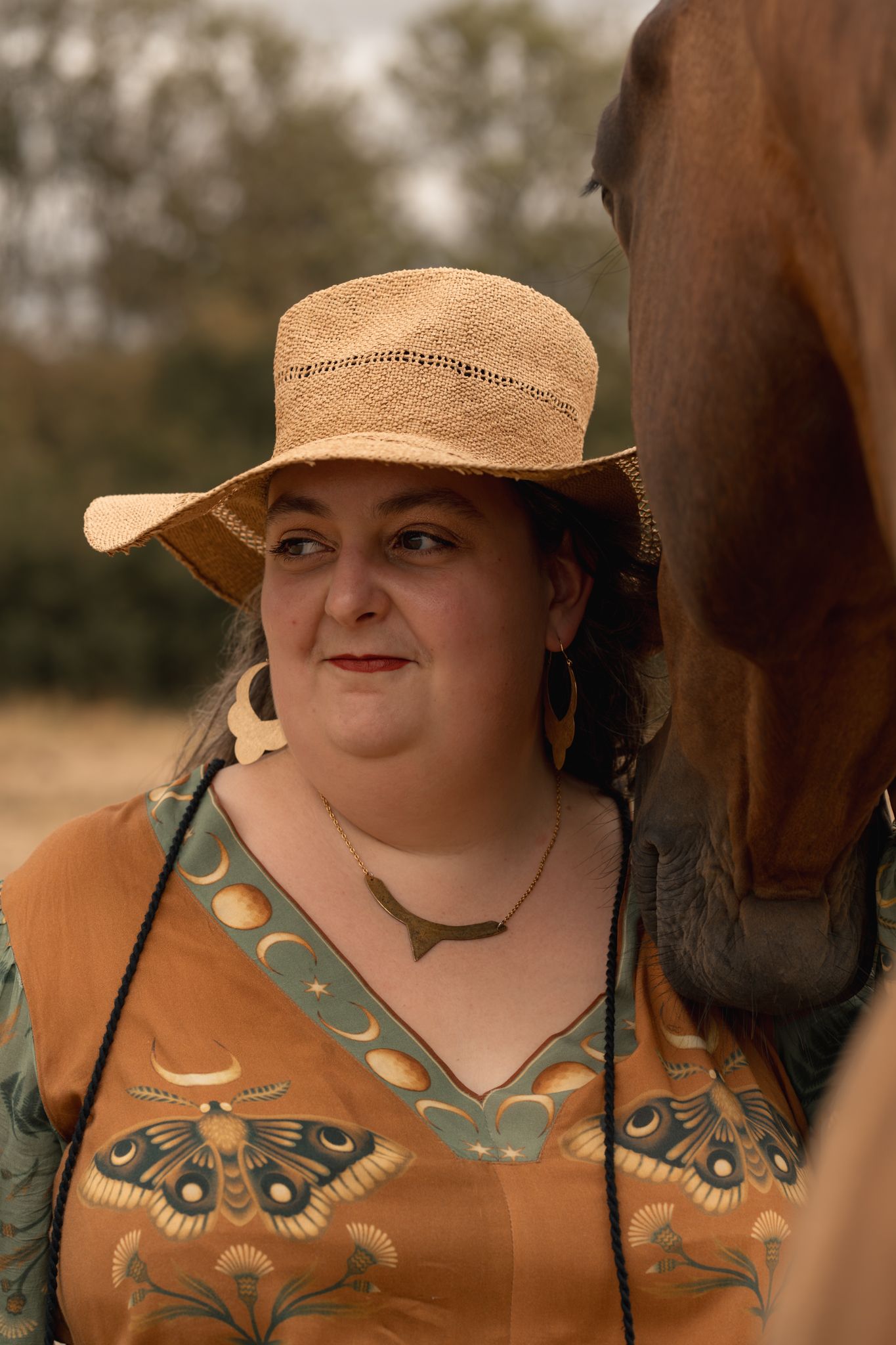 Woman in straw cowboy hat and butterfly print blouse smiling beside a chestnut horse outdoors.
