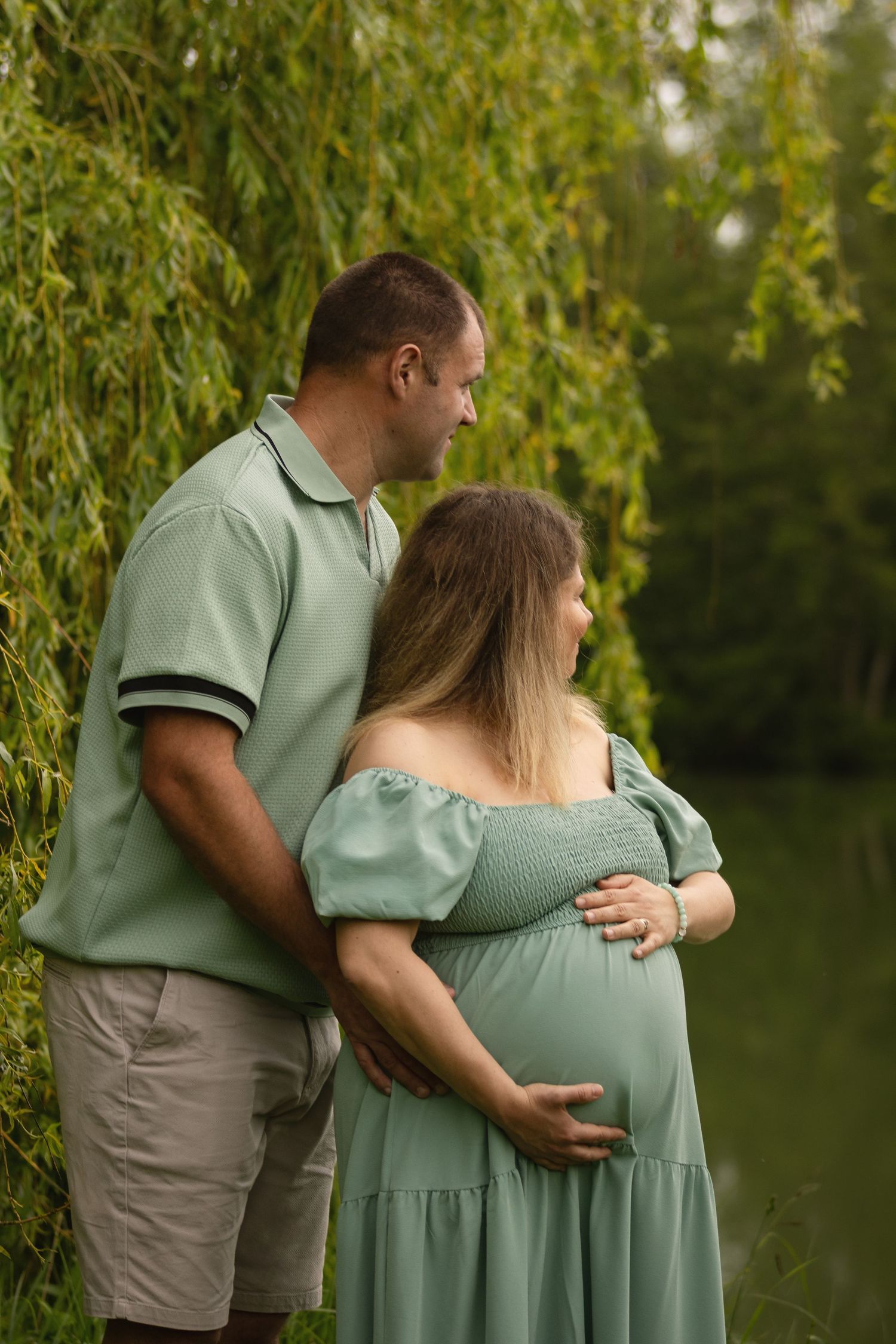 A couple in matching sage green outfits share a tender moment outdoors, the man embracing his pregnant partner near a willow tree.