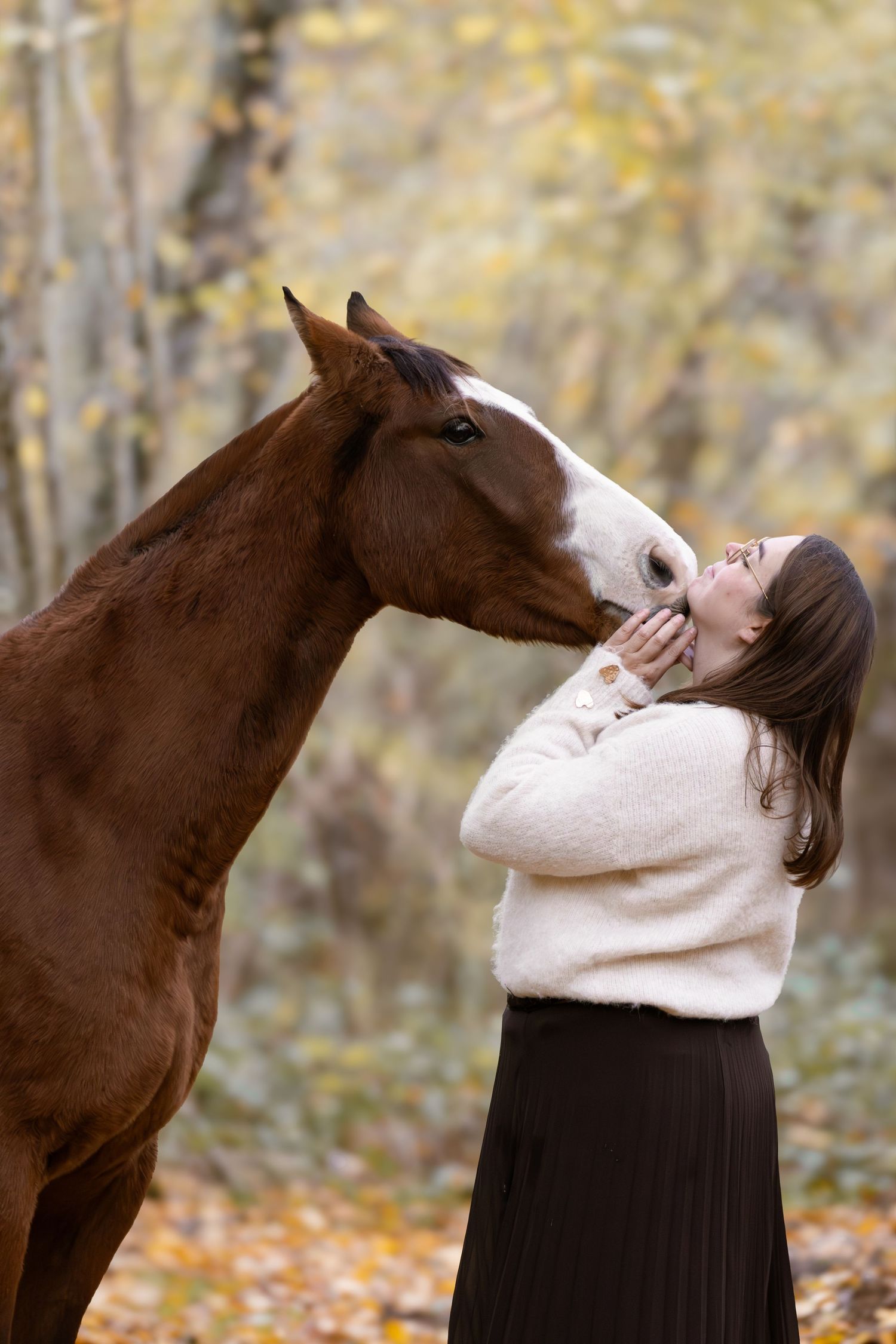 Chestnut horse nuzzling a woman in a white sweater in a colorful autumn forest.