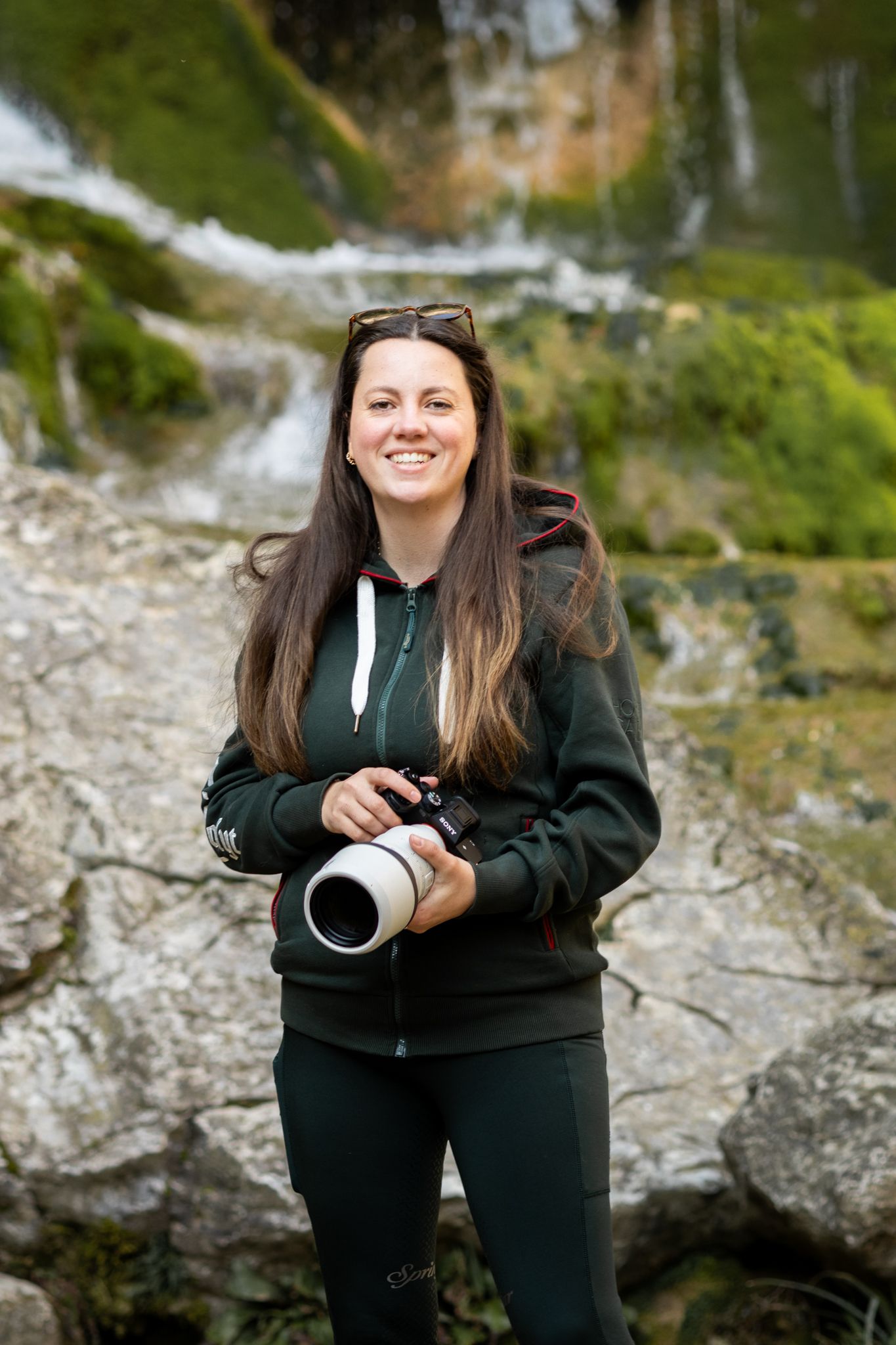 Smiling woman photographer holding a camera with telephoto lens, standing near a waterfall and mossy rocks outdoors.