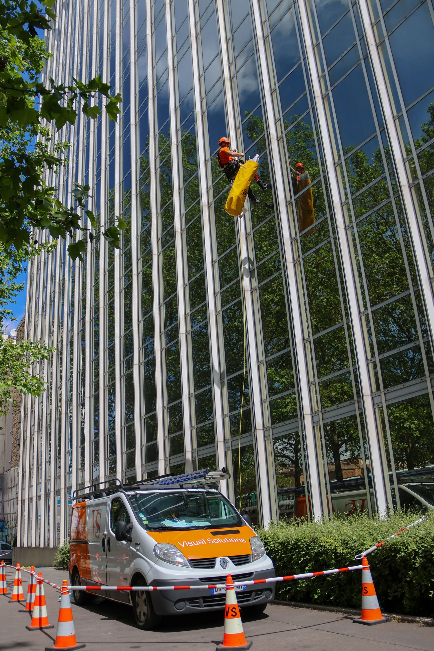 Workers in safety gear clean glass facade of modern building using cherry picker lift truck with orange cones below.