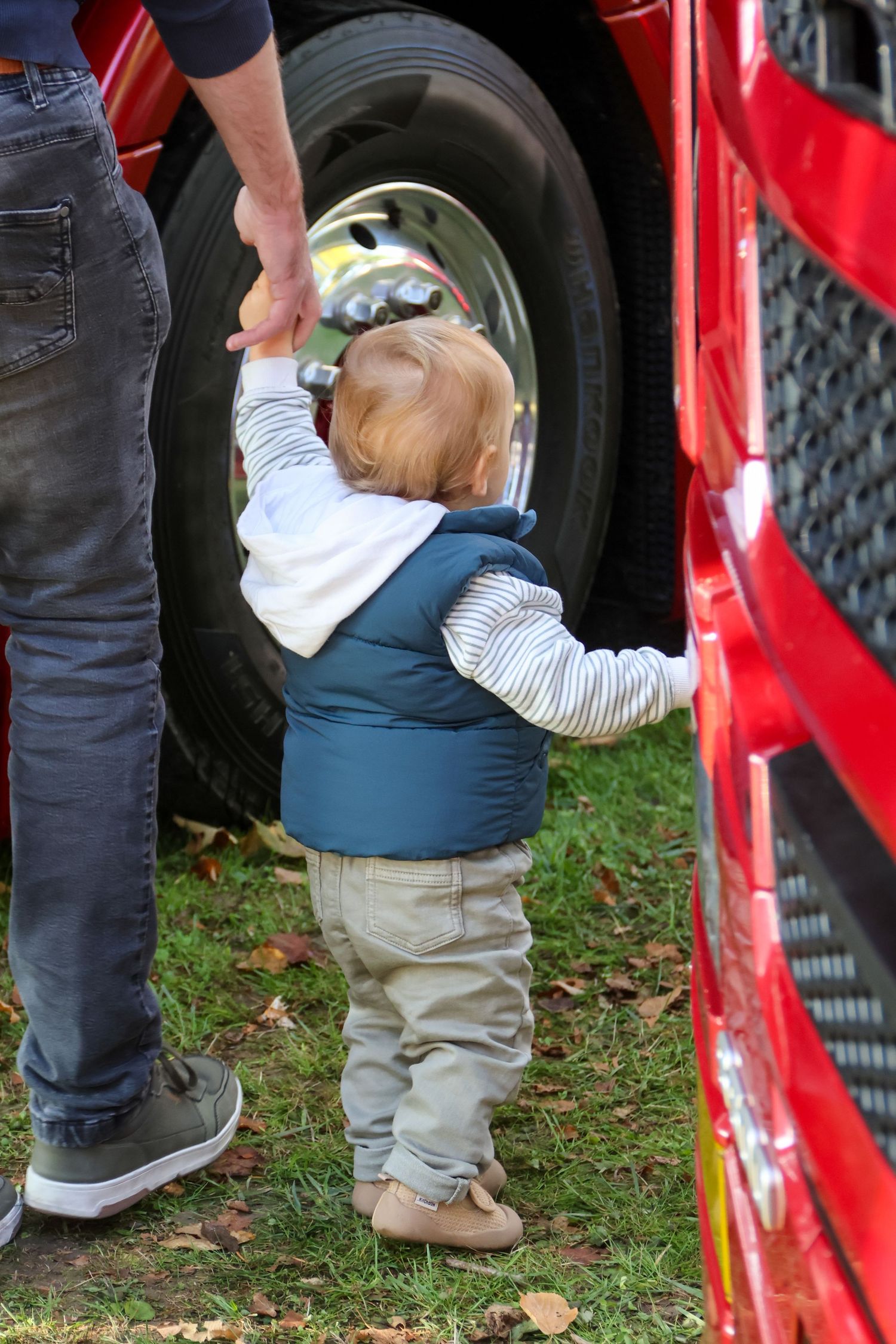 Toddler holding an adults hand reaches toward a shiny red truck tire at an outdoor truck show.