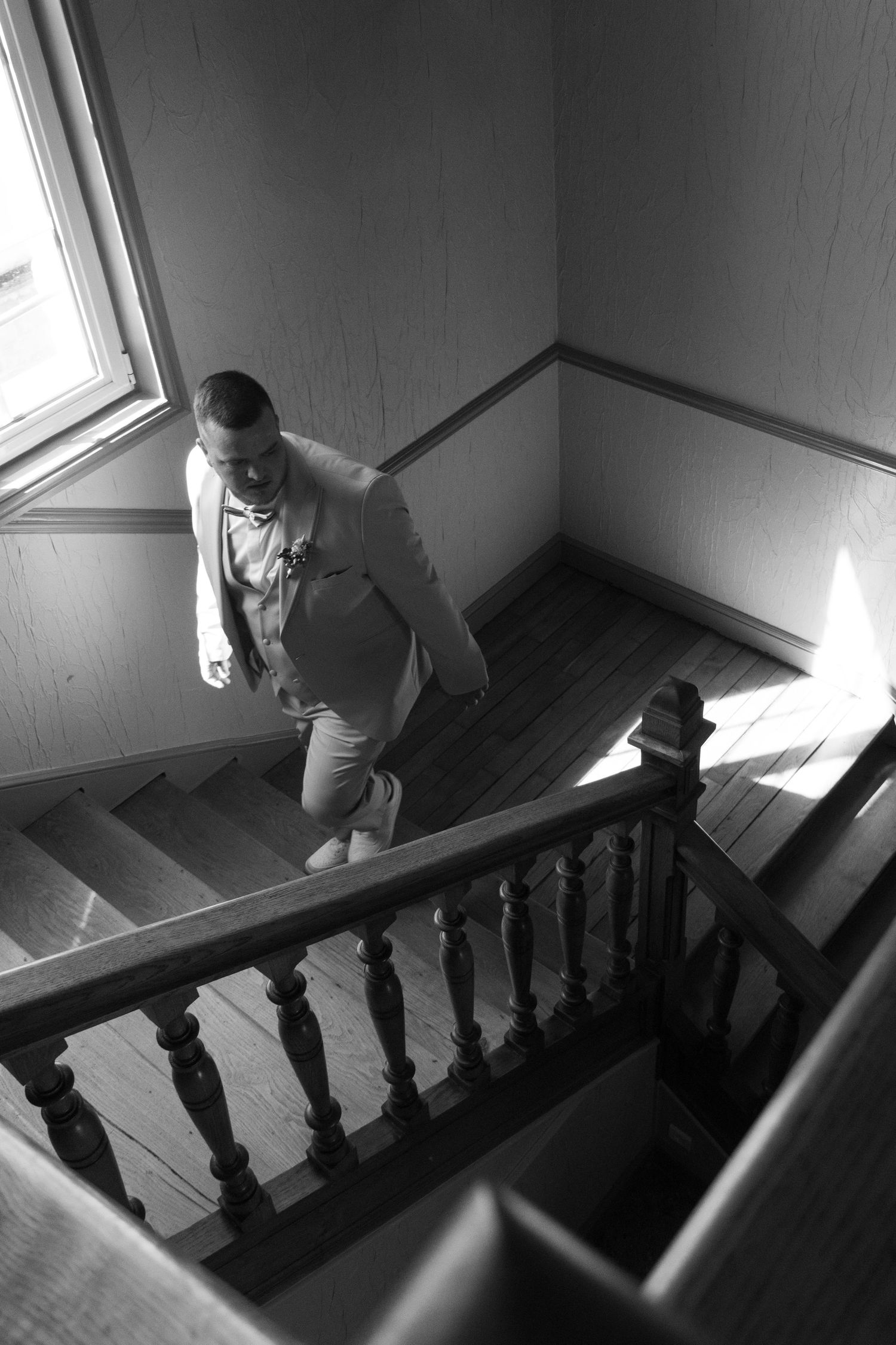 Black and white photo of a man in a suit descending a wooden staircase with ornate balusters near a window.
