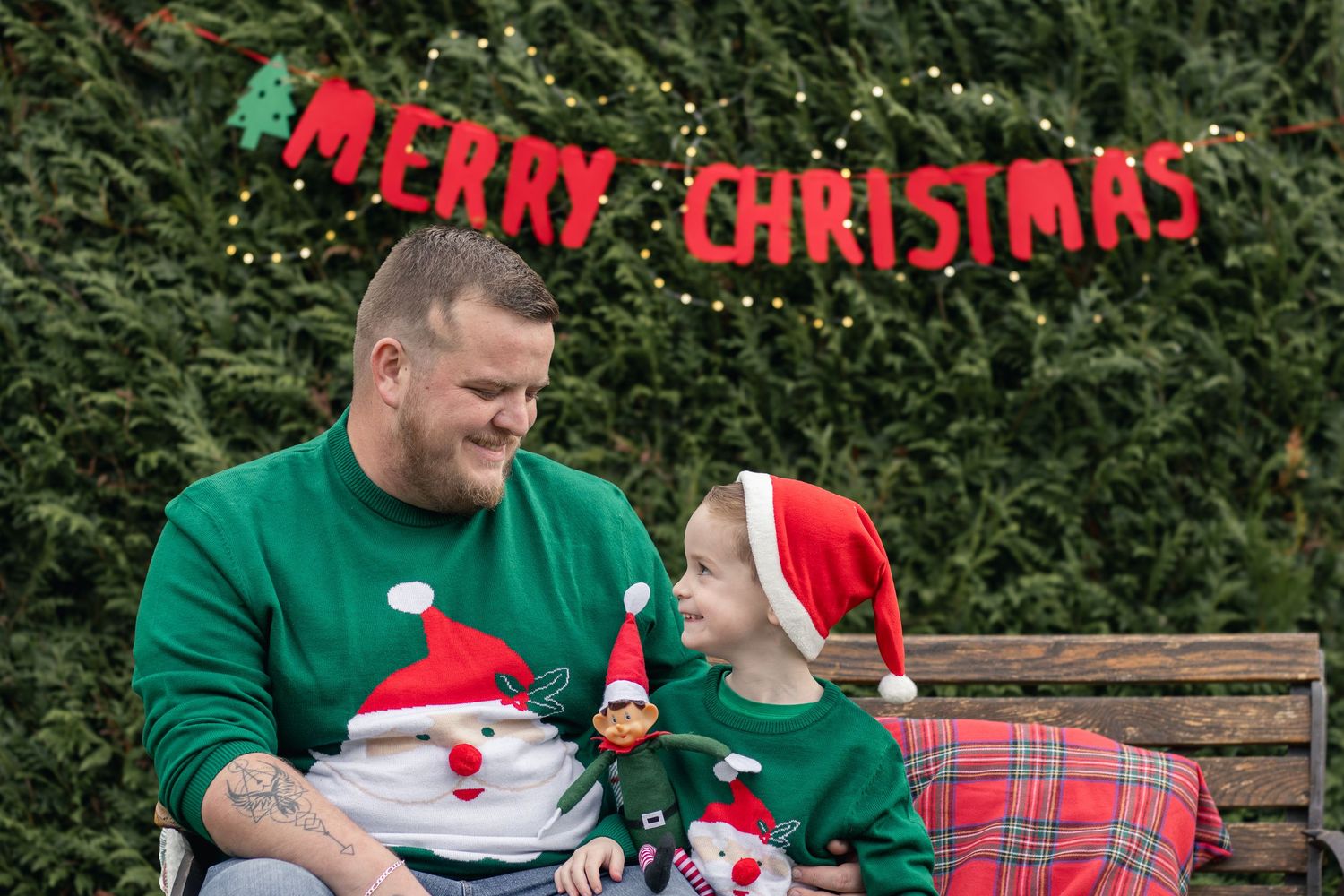 A father and son wear matching Christmas sweaters and Santa hats on a decorated bench.