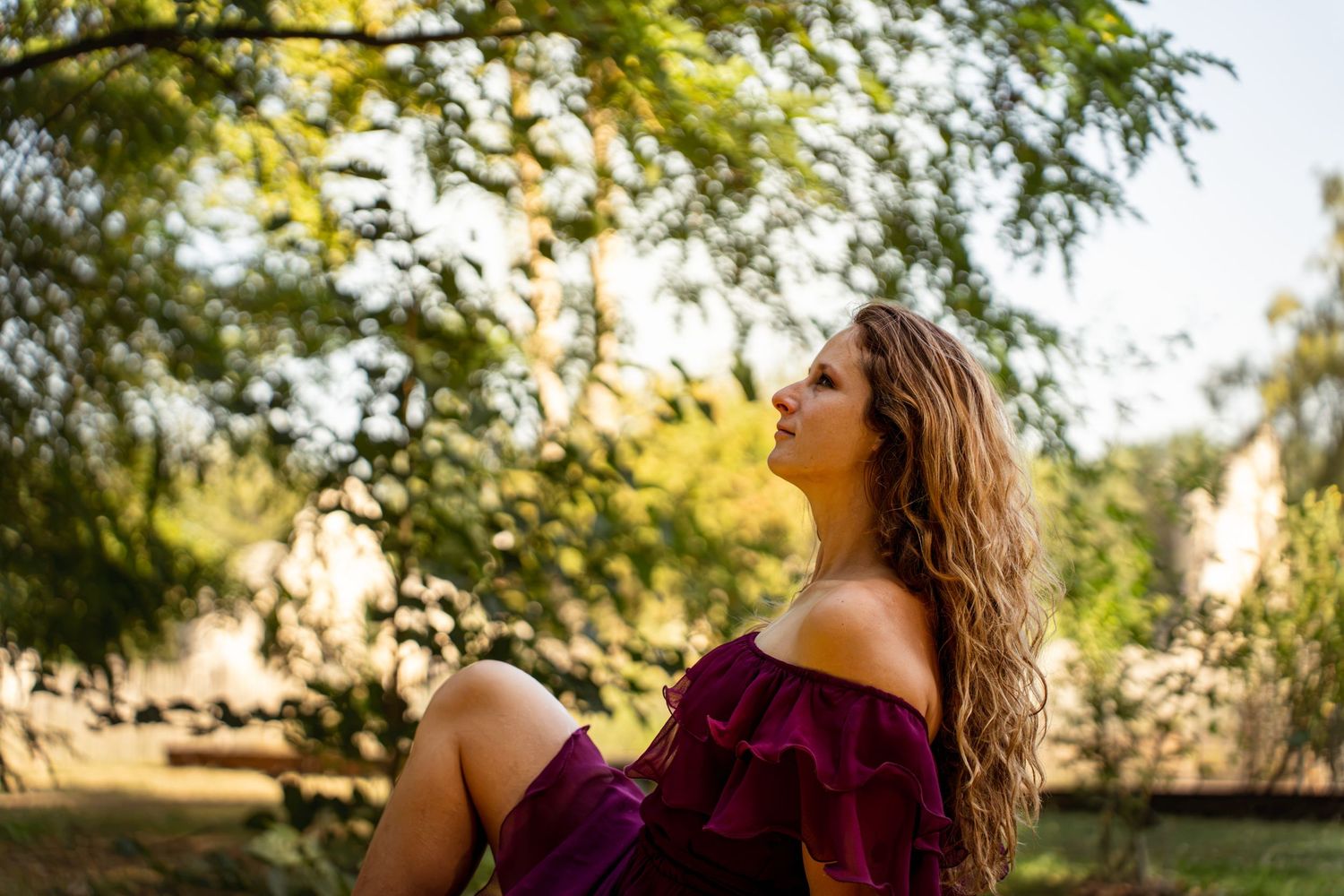 Young woman in purple off-shoulder dress sitting outdoors under lush green trees, enjoying a serene moment in nature.