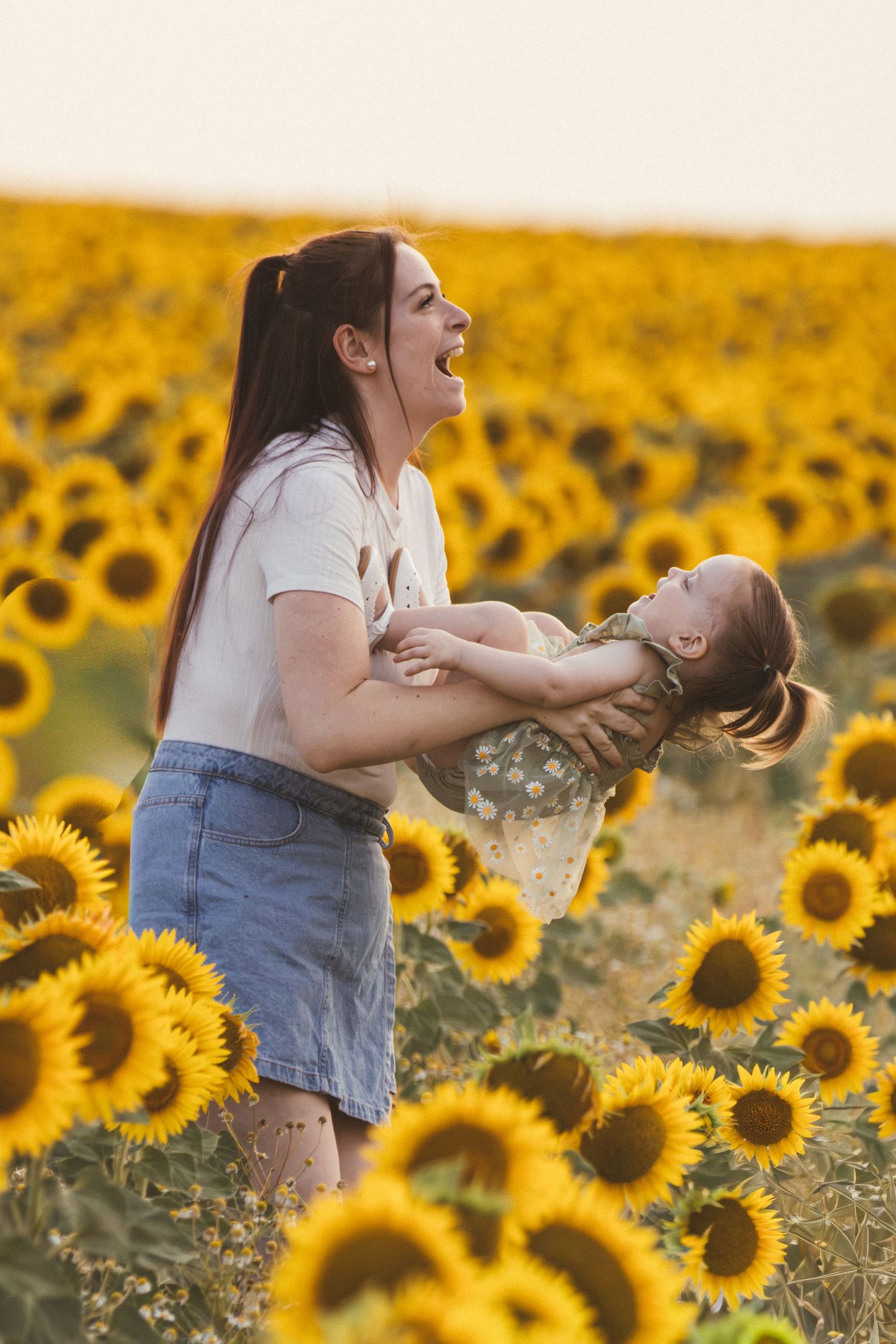 A joyful woman lifts a laughing child in a vibrant sunflower field at golden hour.