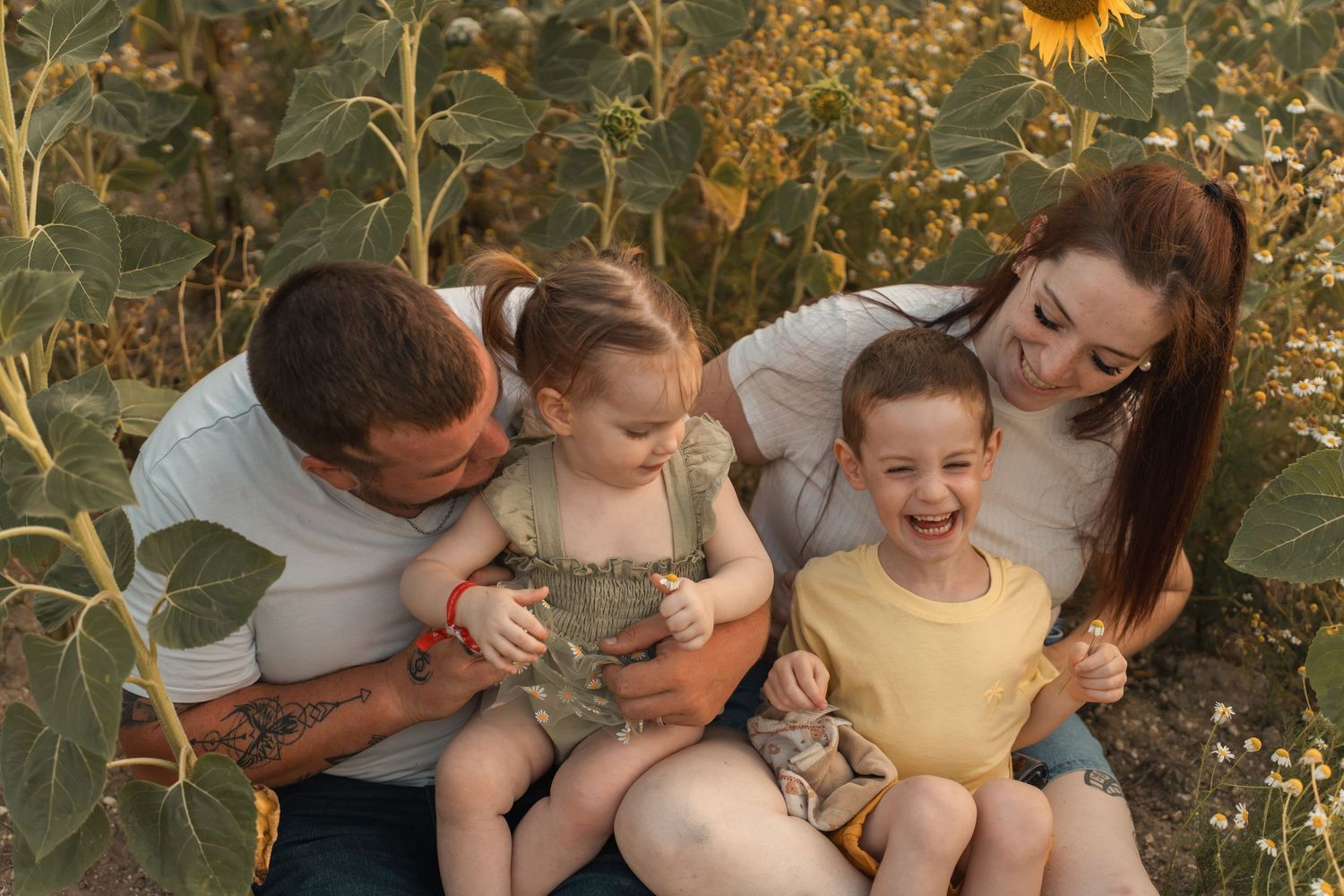 Happy family with two young children sitting and laughing together in a sunflower field on a sunny day.