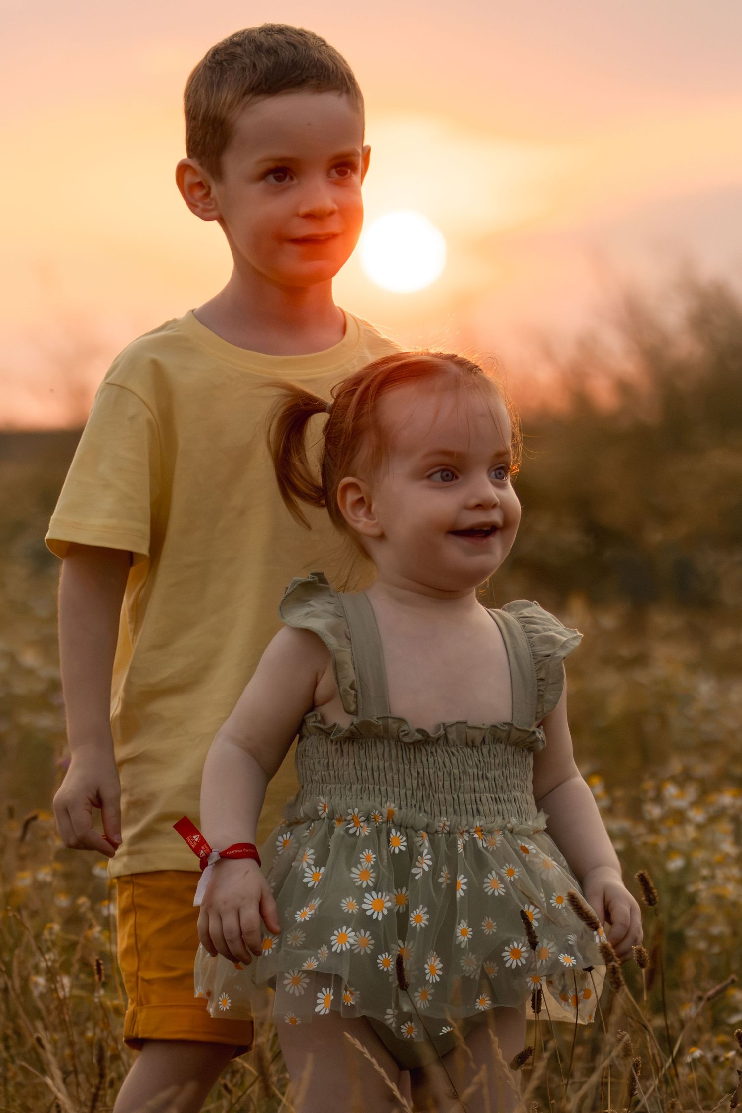 Two children, a boy in yellow shirt and toddler girl in floral dress, stand together in a field at golden sunset.