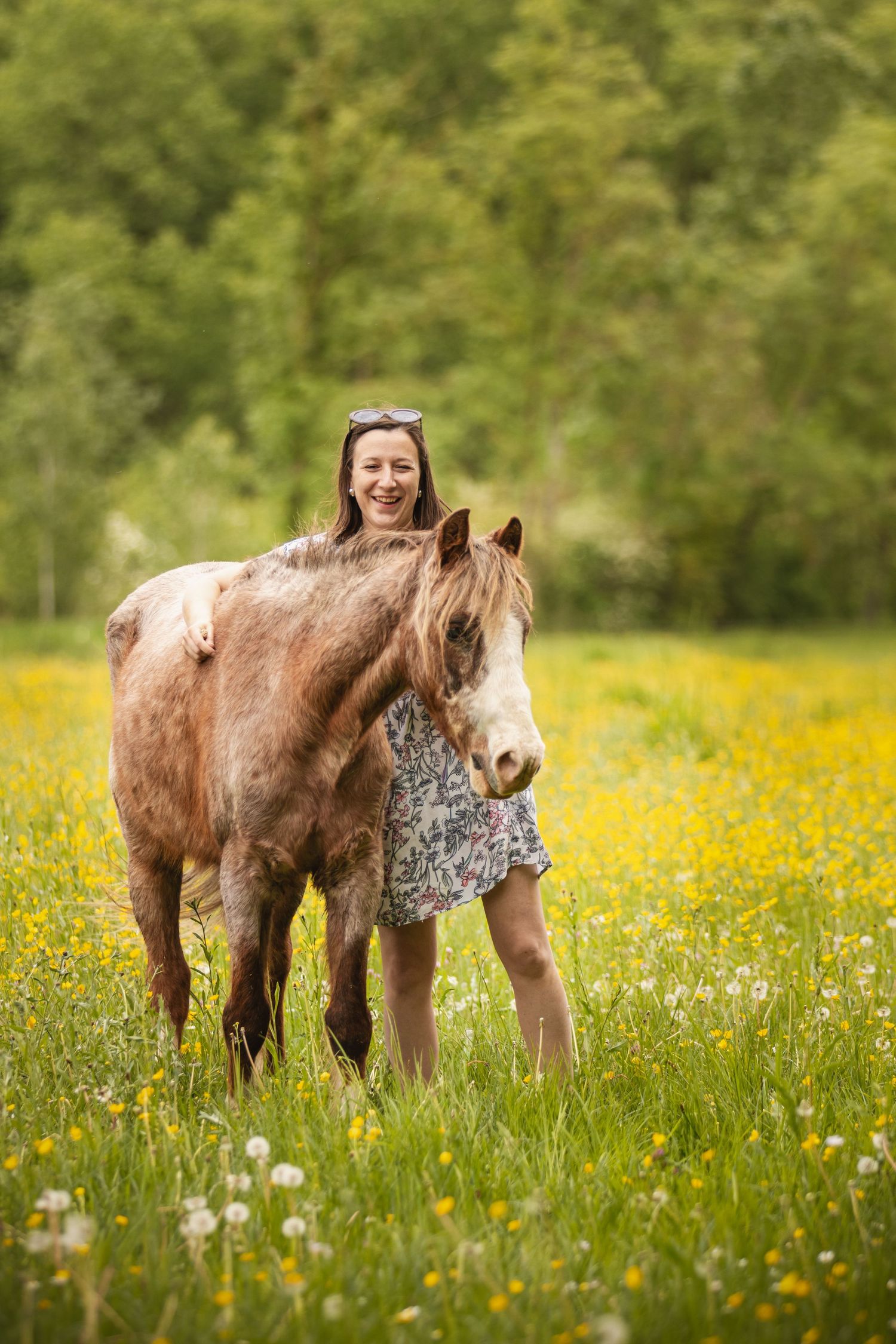 Smiling woman in floral dress standing with a chestnut horse in a sunny meadow filled with yellow wildflowers.