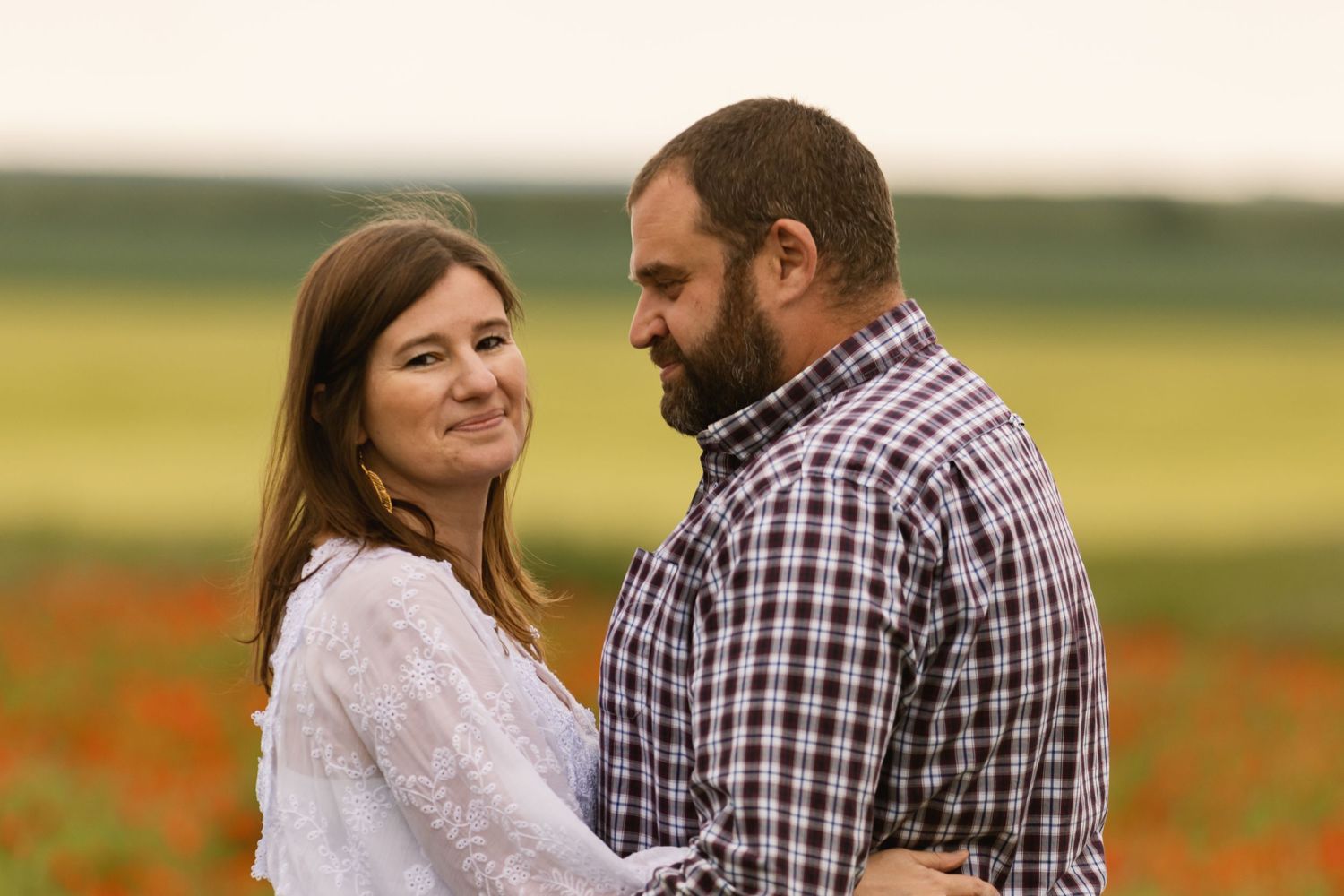 Couple embracing in a blooming poppy field, woman in white dress smiling, man in plaid shirt, romantic outdoor portrait.