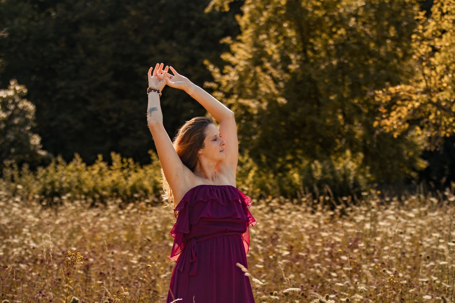 Woman in purple dress raises arms in golden sunlit meadow with wildflowers and trees in background.
