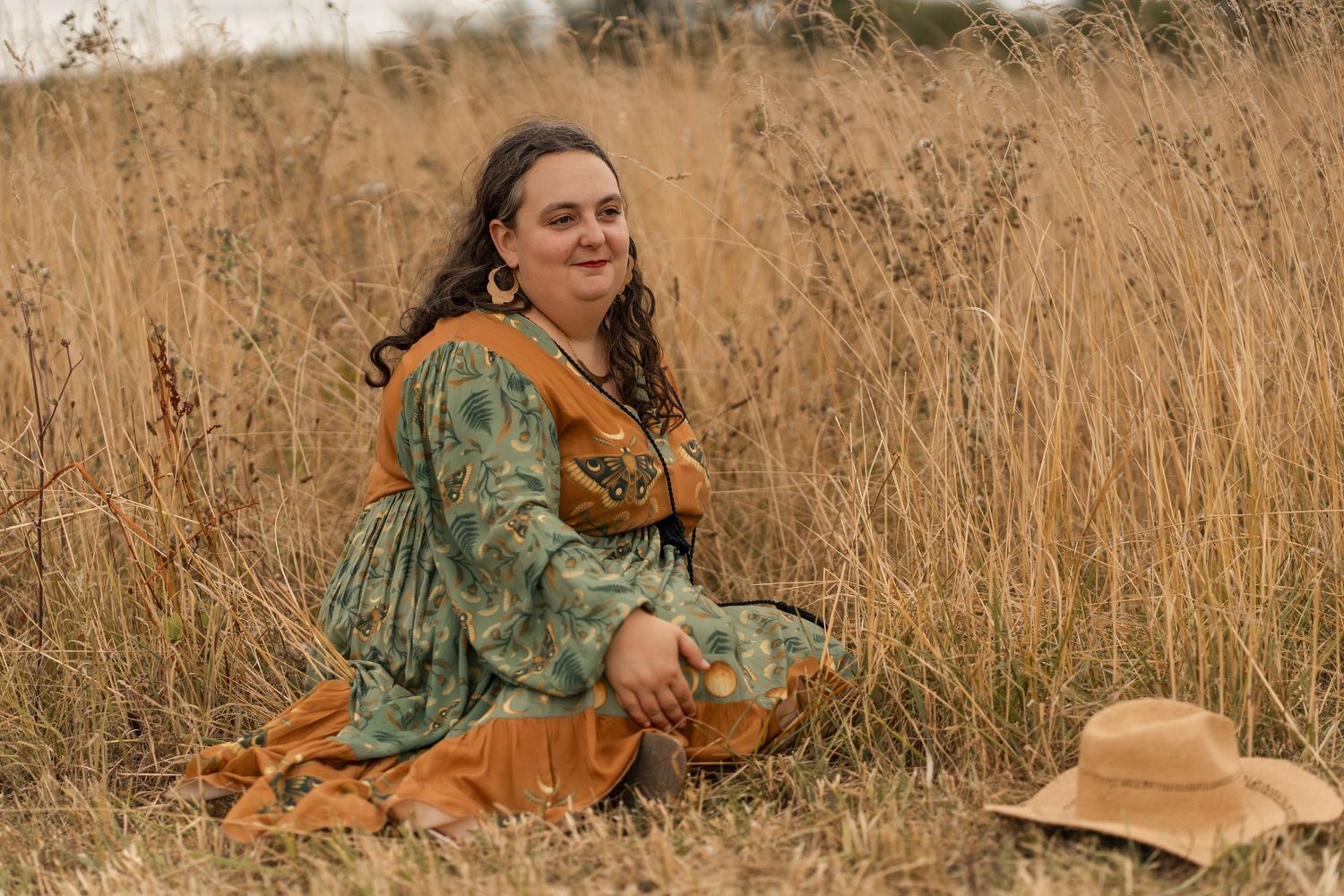 Woman in green floral dress sitting in golden field grass, smiling outdoors.