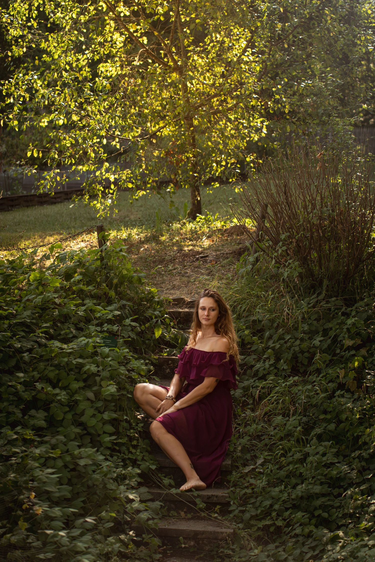 Young woman in burgundy off-shoulder dress sitting on stone steps surrounded by lush green foliage in a sunlit garden.
