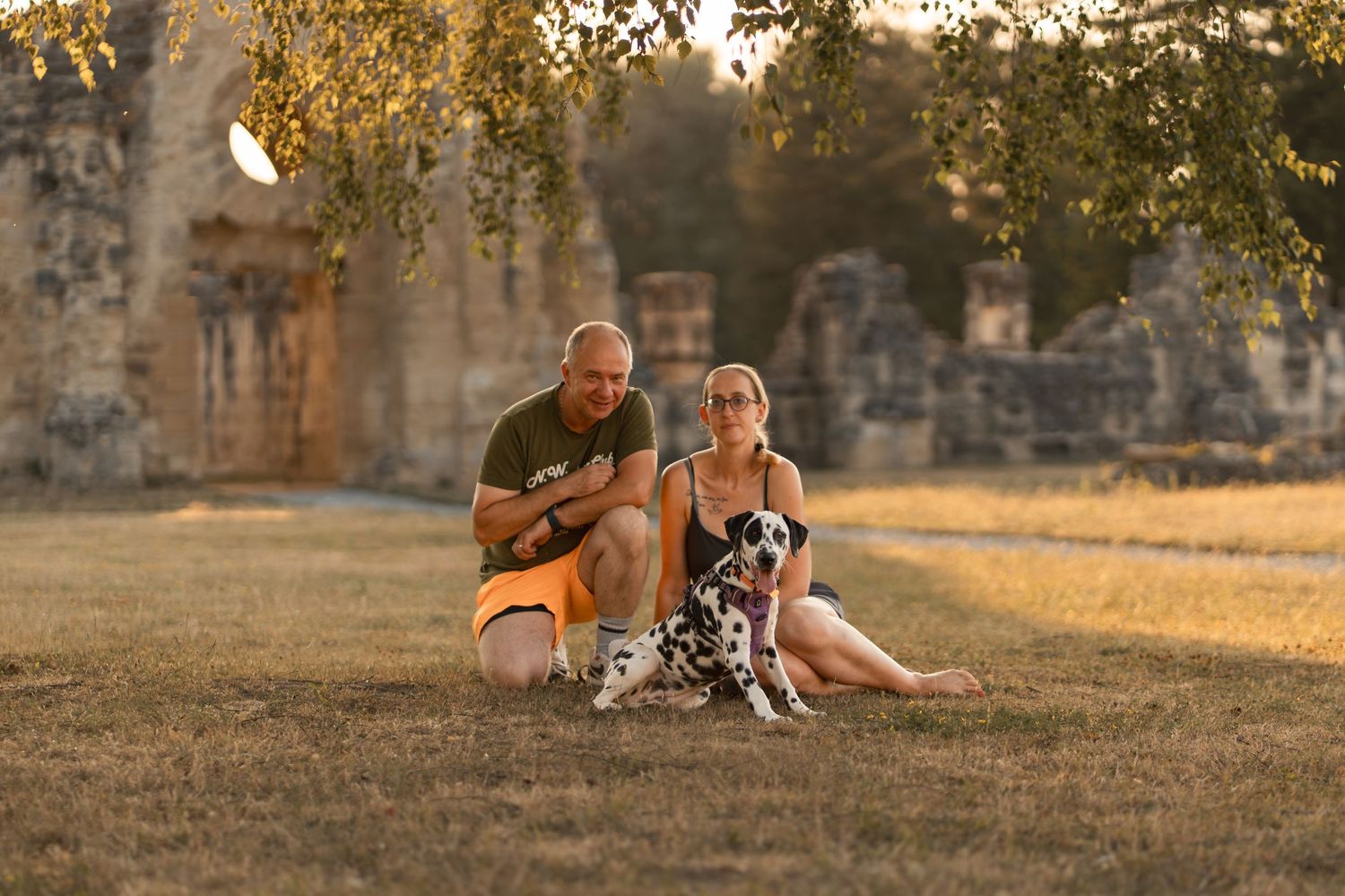 Couple sitting on grass with Dalmatian dog near ancient stone ruins during golden hour sunset.