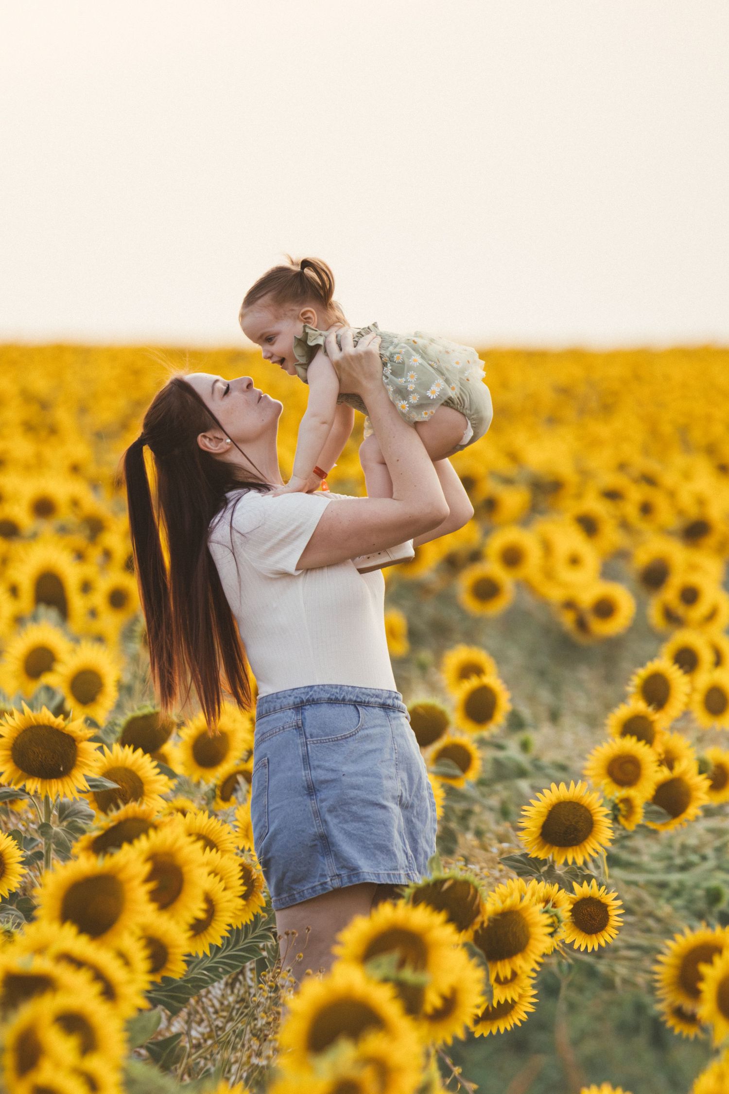 A joyful mother lifts her baby girl in a vibrant sunflower field during golden hour.